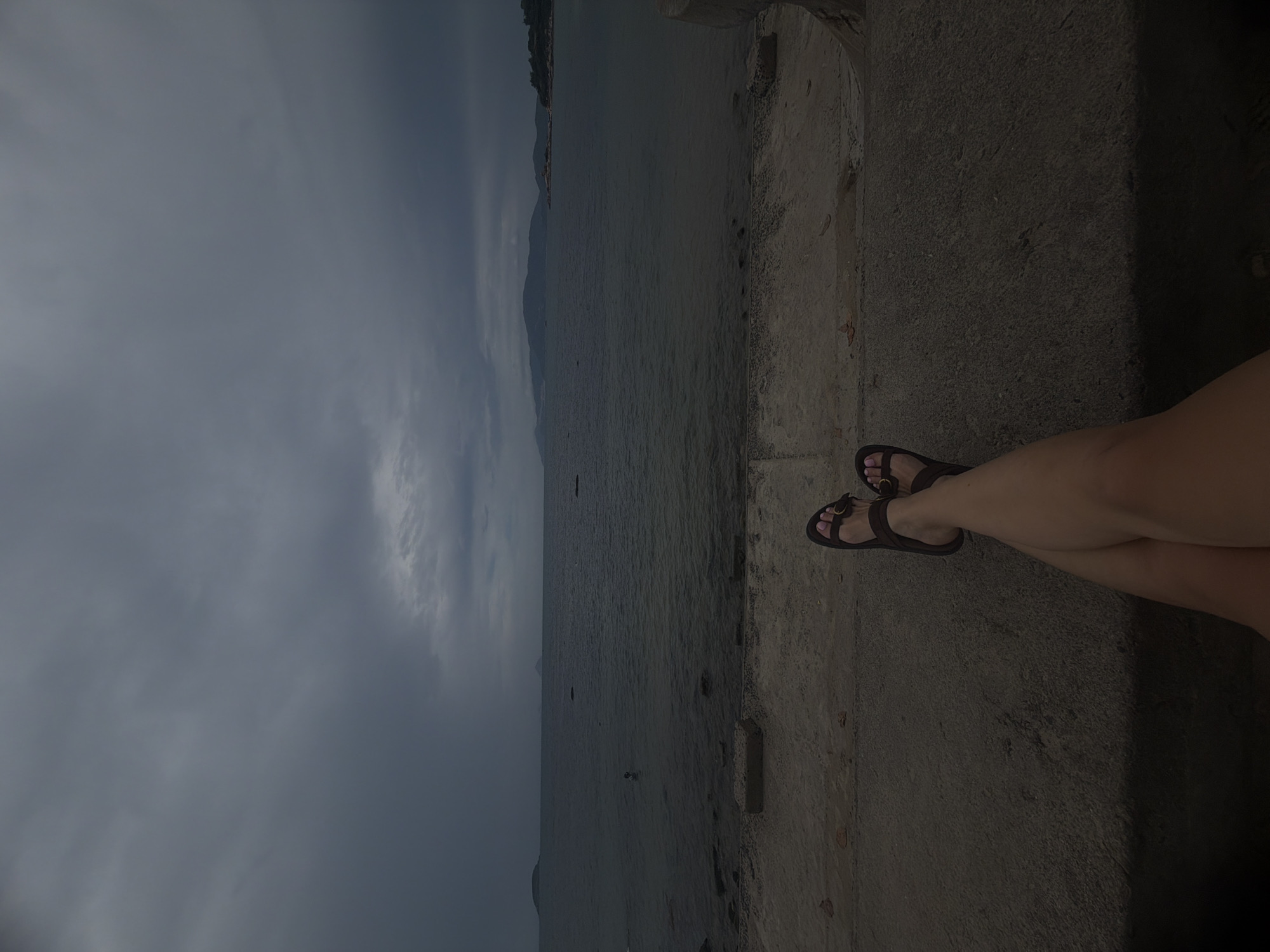 A vertical eye-level shot depicts a cloudy sky above a body of water. On the right side of the frame are the legs of a person wearing brown sandals, resting on a concrete ledge. The scene is presumably set in or near Bac Nha Trang Ward, Vietnam, given the location details provided.