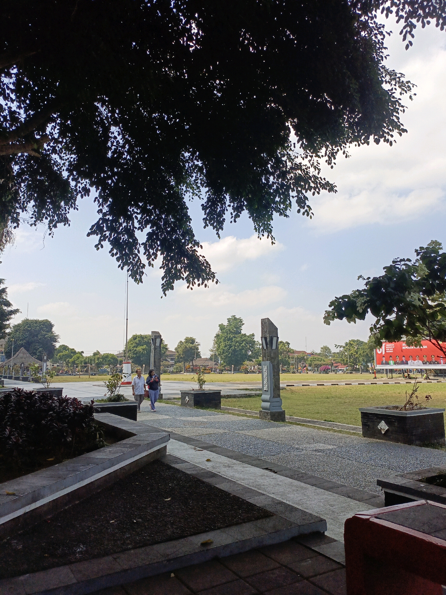 The image depicts an outdoor scene in Wonosobo, Indonesia, likely a public square or park. In the foreground, a patterned walkway leads towards a grassy field. Two people, an adult male and an adult female, are walking away from the camera on the walkway. The male is wearing a white long-sleeved shirt and dark pants, and the female is wearing a dark hooded jacket and light-colored pants. They are walking side-by-side.
Dominating the upper portion of the image is the dark silhouette of dense tree foliage, casting shadows and partially obscuring the bright, cloudy sky. In the mid-ground, a tall flagpole stands prominently in the center of the open space, with a large red banner displaying text and images visible in the far right. Various trees and some low buildings are scattered throughout the background. The overall environment appears to be a well-maintained public area with elements of nature and urban infrastructure.