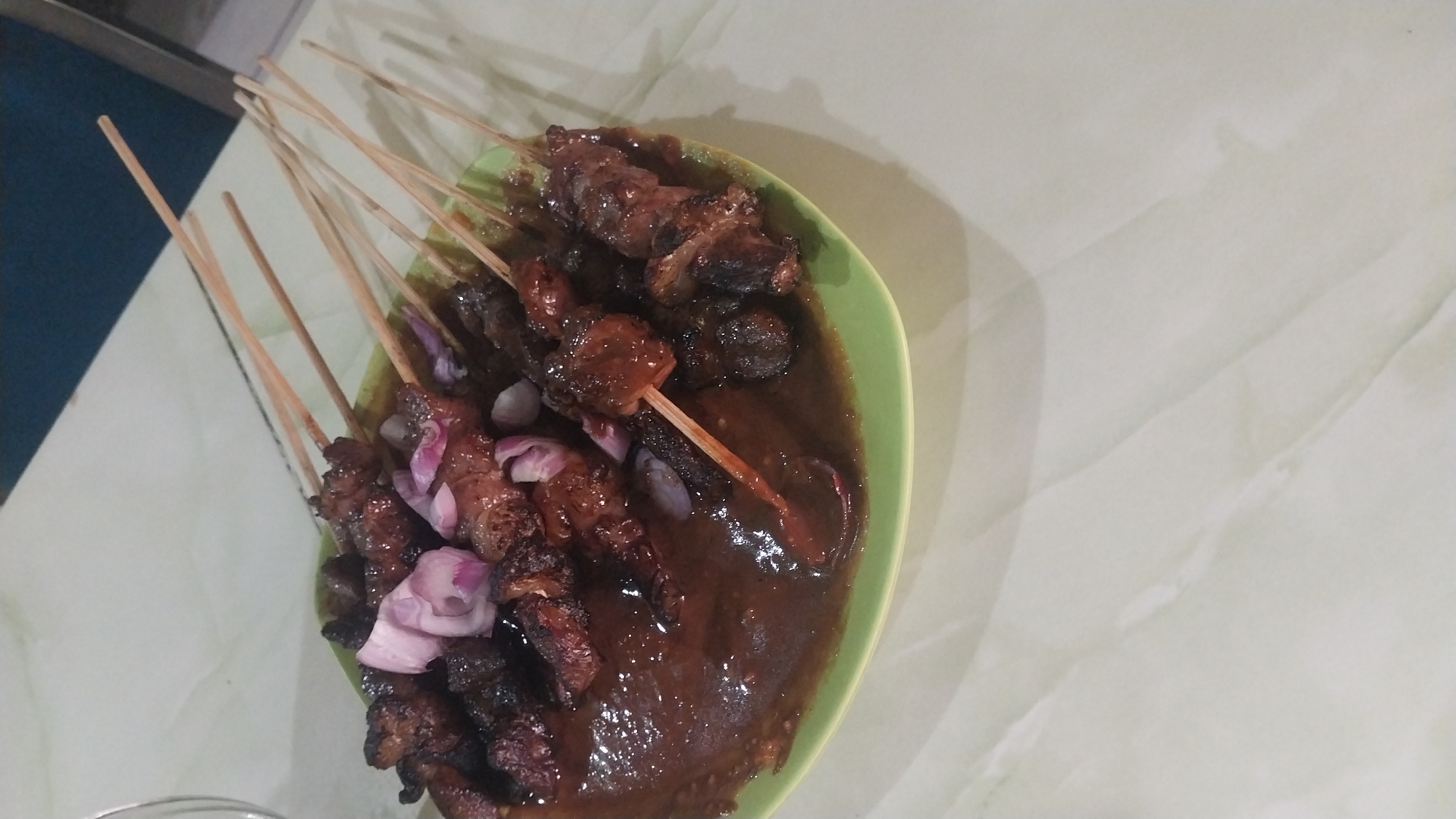 A plate of satay sits on a light-colored, marbled surface. The satay, skewered on wooden sticks, is coated in a dark, rich sauce and garnished with thinly sliced red onions. The green plate is full of the satay, with some skewers extending beyond the plate's rim. The background is a light-colored, textured surface, possibly a table. A dark blue surface is visible in the upper right corner. The lighting suggests an indoor setting, likely a dining area. There are no people visible in the image. The scene is devoid of any explicit geographical markers indicating the city of Gedangan, Indonesia.