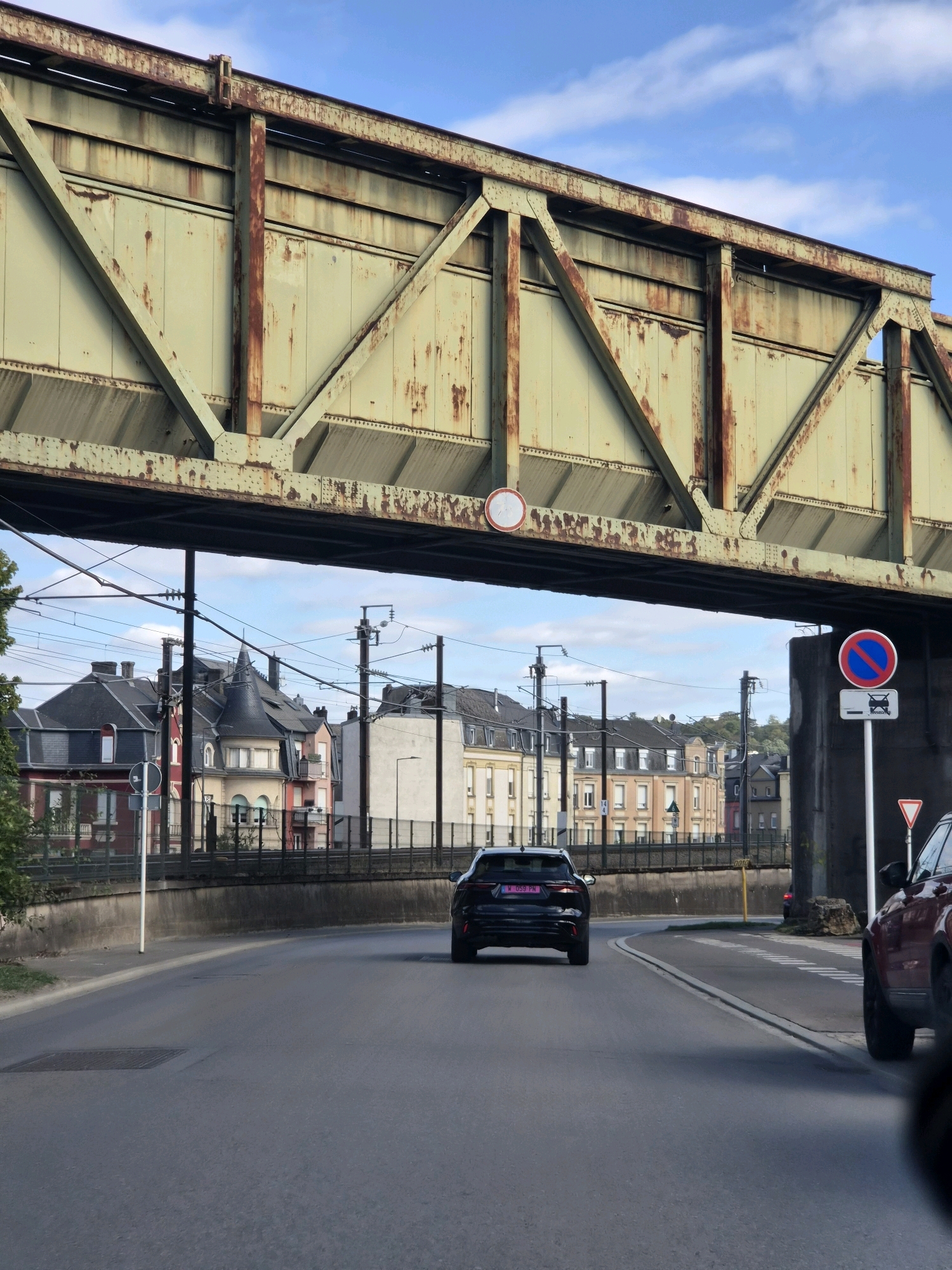 The image shows a road in the city of Esch-sur-Alzette, Luxembourg, with a large, rusty metal bridge overhead. A black SUV is driving away from the camera on the road, with a red vehicle partially visible on the right side. In the background, there are several multi-story buildings with distinctive architectural features, characteristic of European cities. Overhead, there are power lines and traffic signals. A "no entry" traffic sign with a car symbol is visible on a pole next to the bridge. The sky is blue with scattered clouds. The scene appears to be a typical urban environment during daylight hours.