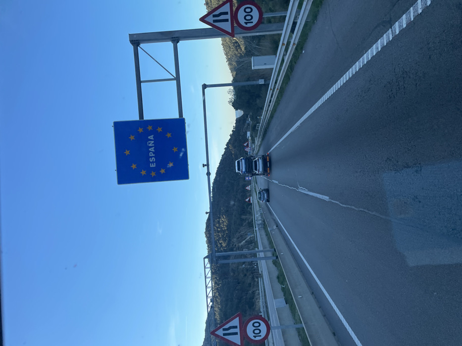 The image depicts a multi-lane asphalt highway on a clear day, identified as a point of entry into Spain near la Jonquera. In the foreground, the perspective is from inside a vehicle, showing a reflection on what appears to be a windshield. The road surface exhibits a prominent longitudinal crack in the center lane.
Dominating the scene above the roadway is a large blue square sign, mounted on an overhead gantry structure, featuring the European Union's circle of twelve yellow stars and the word "ESPAÑA" in yellow capital letters, indicating entry into Spain. Flanking the highway on both sides are road signs: triangular warning signs with converging black lines indicating a narrowing road, positioned above circular white speed limit signs with a red border displaying "100" in black text, signifying a 100 km/h speed limit.
Vehicular traffic includes a car transporter truck in the center lane, carrying at least two white SUVs. Orange cones are visible at the rear of the truck. Ahead of the transporter, a dark-colored passenger car is present in the left lane. Guardrails line both sides of the highway. The background consists of hills covered with dense green vegetation under a clear blue sky.