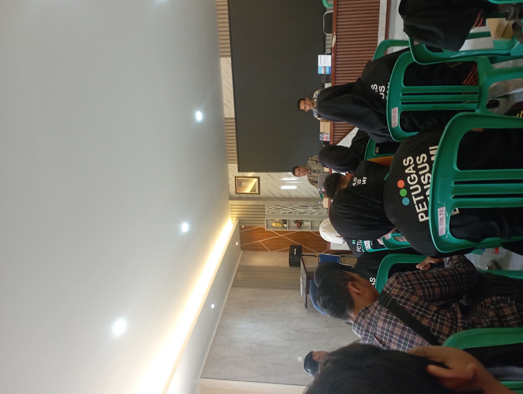 The image depicts an indoor gathering, possibly a meeting or training session, with a group of people seated in green plastic chairs. The attendees are mostly seen from behind, wearing dark clothing, some with hoods. Several individuals have "PETUGAS SENSUS" (Census Officer) written on their backs, indicating the event's purpose.
In the background, two men are seated at a table in front of a large dark screen. One man, wearing a patterned shirt, is looking towards the audience. The other man, in a plaid shirt, is also seated at the table, facing forward. The room has modern decor, with textured walls and strip lighting on the ceiling. To the right of the screen, a tall, slender shelving unit displays awards and decorative items. A podium-like structure with a table is visible on the left side of the room.
The overall lighting suggests a well-lit indoor space, with natural light possibly coming from the left, indicated by the illuminated ceiling. The presence of a large screen and seating arrangement points to a presentation or instructional setting. While the image does not directly provide evidence of its specific location within Garut, Indonesia, the text on the shirts suggests a connection to official census activities in the region.