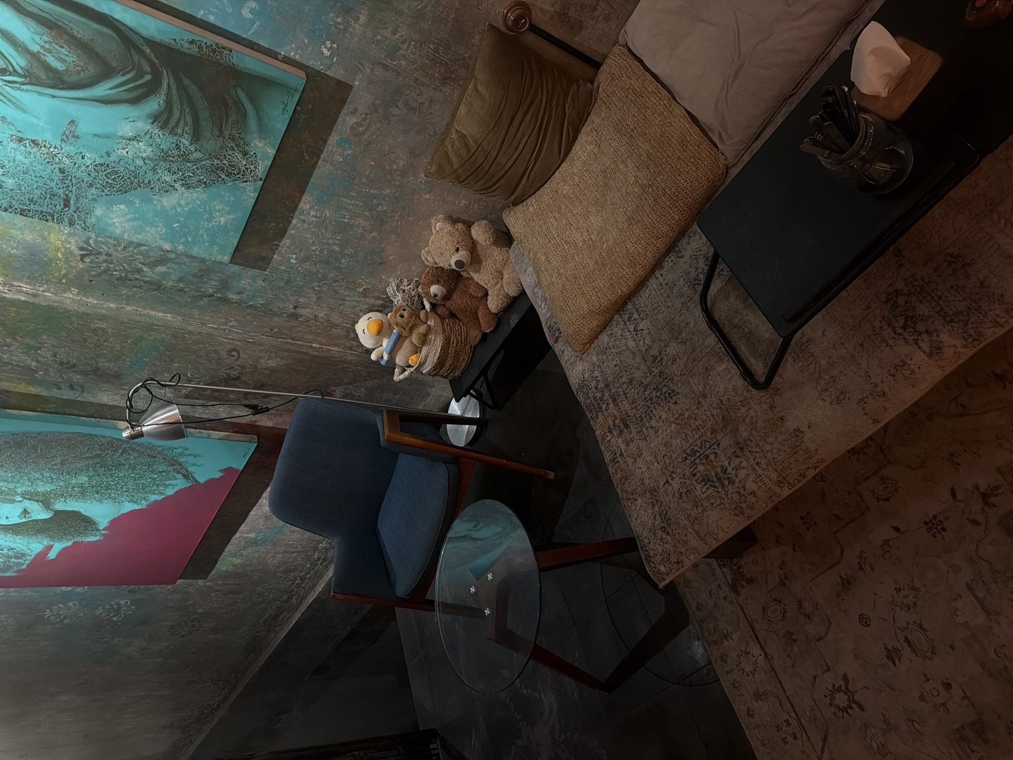 The image depicts an indoor scene, possibly a seating area within a cafe or a lounge, in Ubud District, Indonesia. The viewpoint is from an elevated angle. On the left, there is a piece of abstract art on the wall, featuring shades of blue and pink. A flexible lamp is attached to the wall near the artwork. In the lower-left, a blue upholstered chair sits near a round glass-topped table with a dark wooden frame. To the right, a sofa is seen with a combination of throw pillows, including a beige cushion and a textured brown pillow. Teddy bears and other stuffed animals are grouped together near the sofa. A small black table is next to the sofa with a glass jar filled with utensils and a stack of napkins. Beneath the sofa and the chairs, an area rug with a faded pattern covers the floor. The room's walls and floors feature a textured, mottled surface, lending a rustic feel to the space.