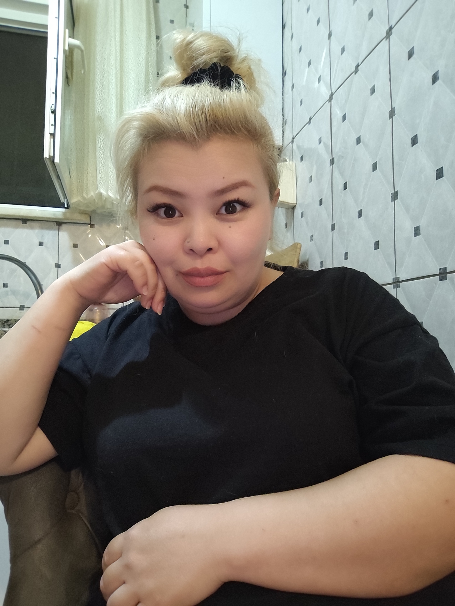 A woman with blonde hair pulled into a bun is seated, wearing a black t-shirt. Her left arm is resting on an olive-green chair, and her head is propped up by her left hand. The background shows a window with a white frame and a sheer curtain, as well as a tiled wall with a geometric pattern of white tiles with small black squares. A faucet is also visible near the window. The location is likely indoors and inferred to be Sarıyer, Turkey. No further details are provided about the subjects, objects, actions, interactions, or environment.