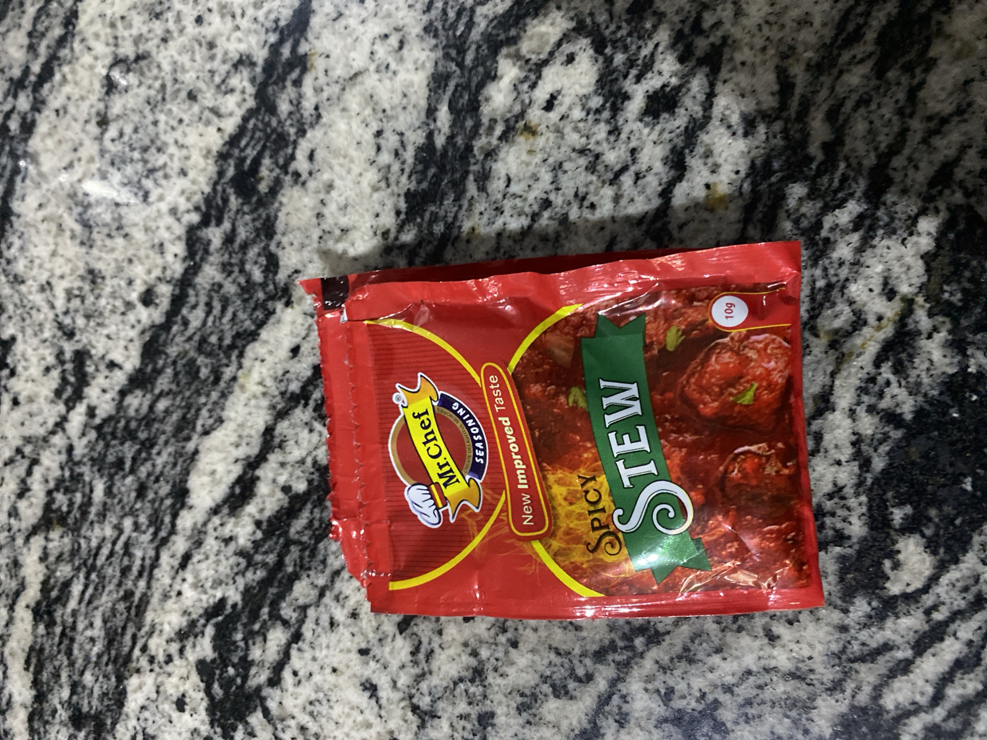 A red packet of Mr. Chef Spicy Stew seasoning is placed on a grey, speckled countertop. The packet features a cartoon chef logo, text indicating "New Improved Taste," and the words "SPICY STEW" in large green lettering. A small red circle with "10g" is visible on the bottom right corner of the packet. The background is out of focus, showing the texture of the countertop. There is no indication of the city of Bar Jirgi Summa, Nigeria in the image.