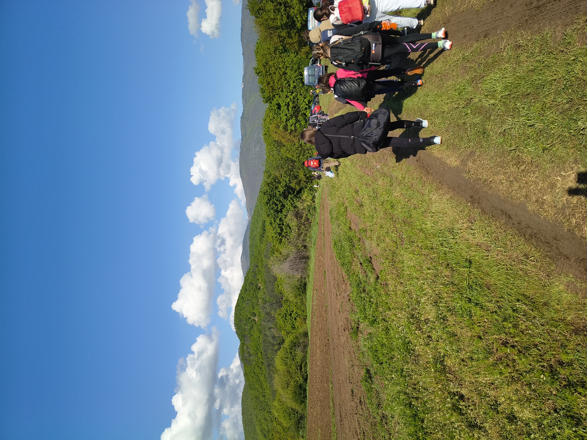 A slightly tilted outdoor shot shows a group of people walking or standing on a dirt path through a grassy field. A mountain range covered in trees is visible to the left. Above the mountain, there's a blue sky with scattered white clouds.
The location cannot be confirmed as Topolovo, Bulgaria, with certainty based on visual information alone.