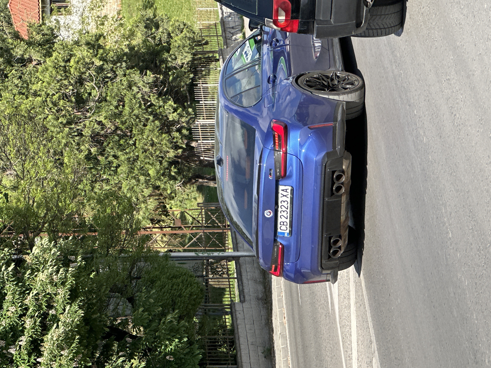 A blue BMW M5 with Bulgarian license plate "CB 2323 XA" is parked on the side of a road, facing away from the camera. The car has quad exhaust pipes and custom rims. A black SUV is partially visible in front of the BMW. A green hedge and trees are on the left side of the image, behind a low metal fence. The scene appears to be in an urban or suburban area, with houses visible in the background. The road is grey asphalt with white lane markings. The location is likely Veliko Tarnovo, Bulgaria, based on the Bulgarian license plate.