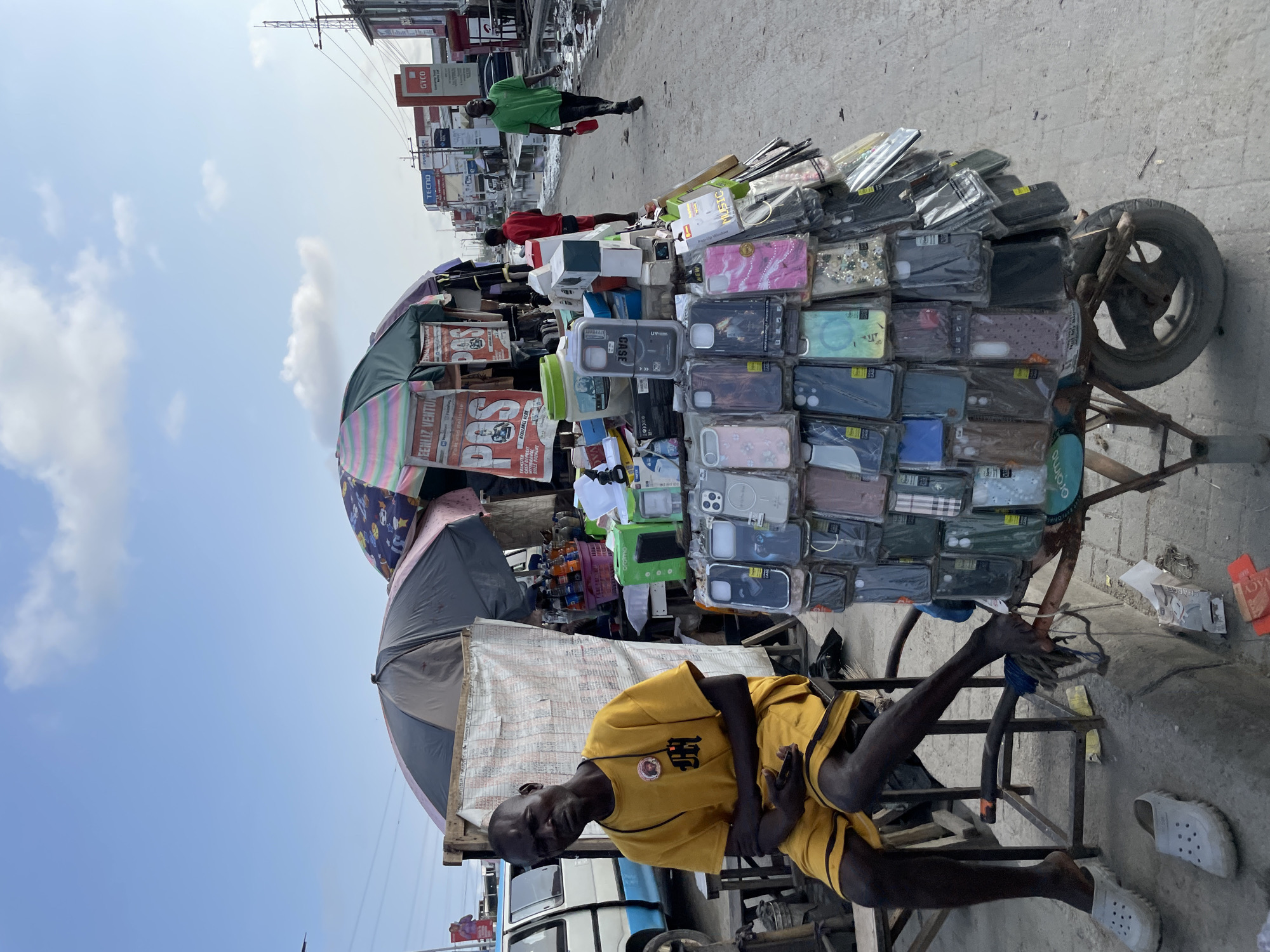 A man in a yellow t-shirt and shorts sits on a chair at a street stall in Lekki, Nigeria. He is looking towards the camera with a neutral expression. The stall is laden with many mobile phone cases of various designs and colors, displayed on a cart. Behind the man, there are two umbrellas, one dark grey and the other a multi-colored patterned one. A white van is partially visible to the left of the frame. In the background, other stalls and shops line the street, with several people walking along the pavement, some carrying items. The street appears wet in places, suggesting recent rain. The sky is partly cloudy with a clear blue expanse.