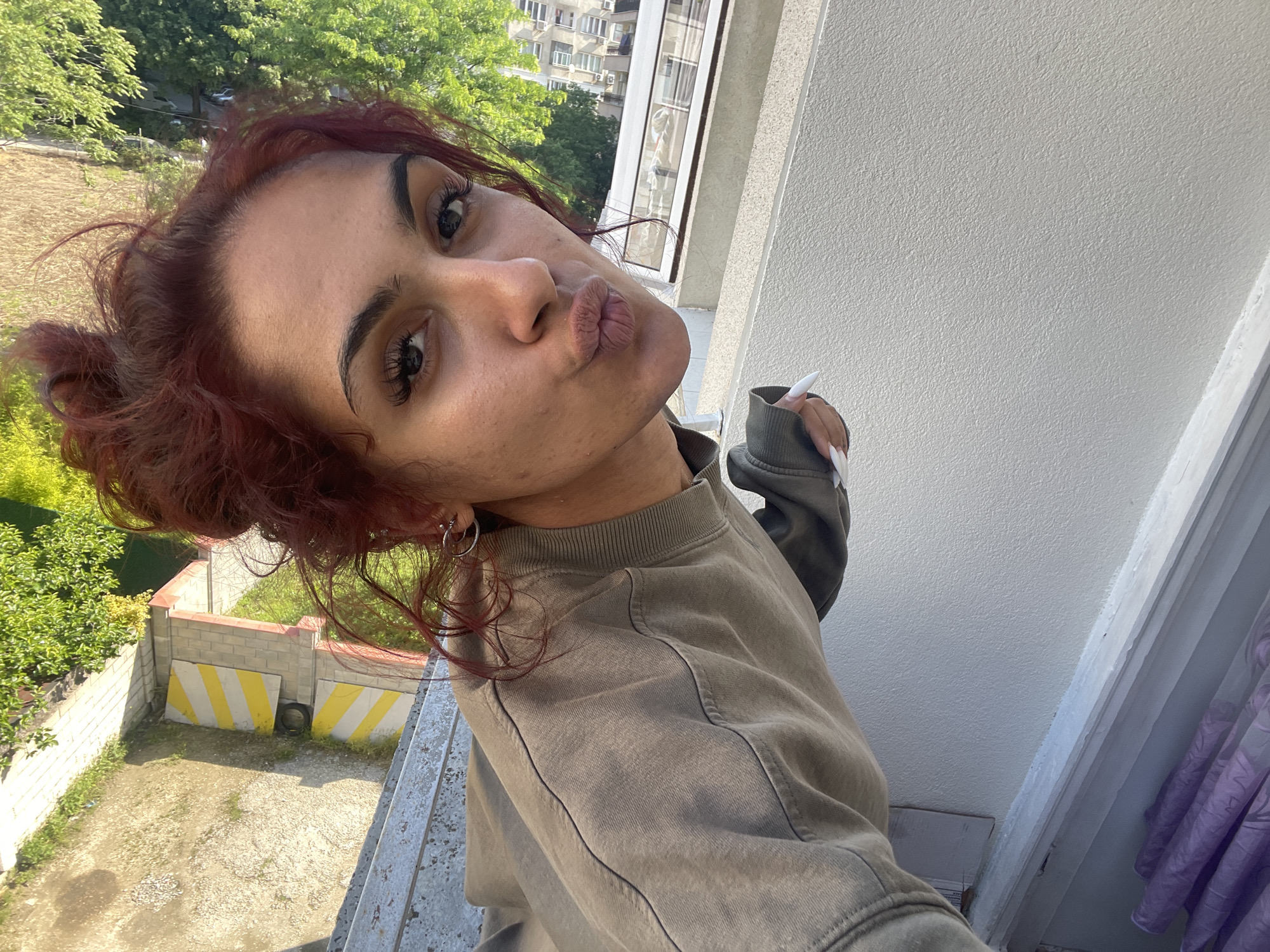 A young individual, positioned on a balcony, is captured in a close-up, tilted selfie. The person has reddish-brown hair, dark eyebrows, and prominent eyelashes, and is pouting their lips towards the camera. They wear a muted olive-green or khaki-colored sweatshirt with distinct seam details and silver hoop earrings. Their right hand, adorned with long, pointed white nails, is partially visible, holding a small white object. The background features a sunlit residential area: to the left, green trees and distant buildings are visible above a lower-level patch of ground with sparse grass, dirt, a low concrete block wall, and two yellow-and-white striped barriers. To the right, a textured white building facade and a window with a sheer, light-colored curtain are present, alongside a multi-story apartment building in the distance. A concrete or metal balcony railing is visible in the foreground. The scene is brightly illuminated by natural sunlight in the city of Unknown, Unknown.