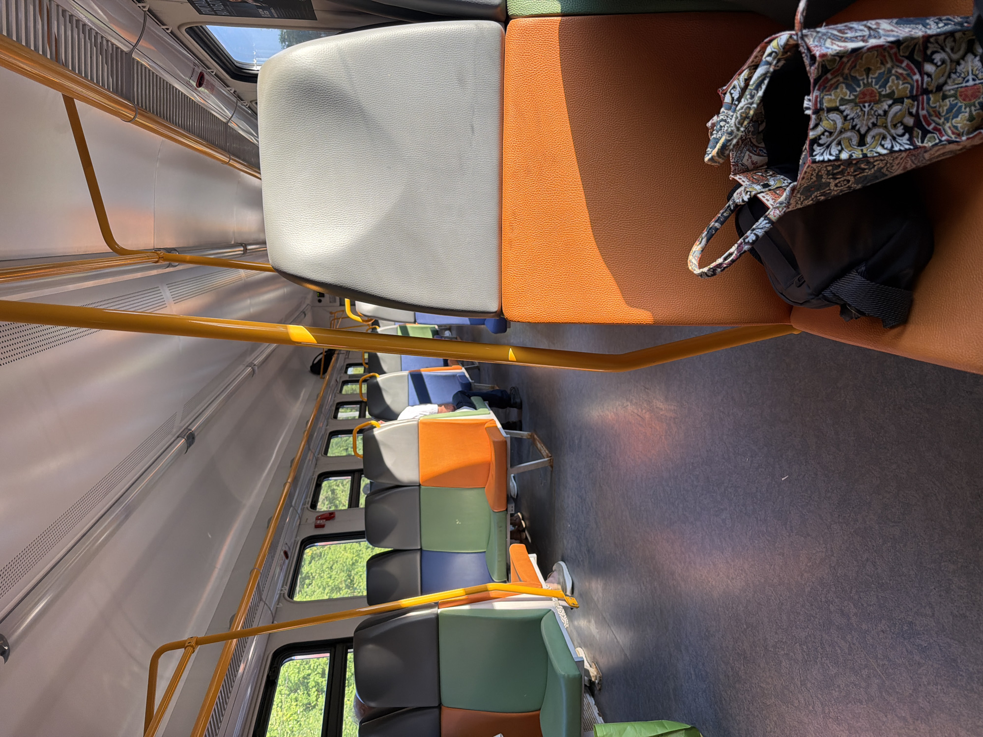 The image shows the interior of a train car, likely in Voskresensk, Russia, during daylight hours. The train car features rows of multi-colored seats, with sections in grey, blue, green, and orange. Yellow handrails are visible along the ceiling and on the backs of some seats. Sunlight streams in through the windows, casting long shadows on the floor and highlighting the textures of the seats. In the foreground, on an orange seat, rests a patterned tote bag and a black backpack. A person can be seen in the background, seated further down the aisle. The overall environment suggests a public transportation setting, possibly during a journey.