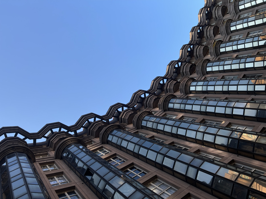 A low-angle, upward-looking photograph captures the monumental, postmodern architecture of the Palacio d'Abraxas in Noisy-le-Grand, France, against a clear, azure sky. The primary subject is a multi-story building facade, composed of alternating sections of light-brown concrete and dark, reflective glass panels that form vertical, rounded column-like structures. Above these glass sections, dark grey, pre-stressed concrete elements create a distinctive wavy, tiered pattern, resembling interconnected, sculptural balconies or brise-soleil. The building extends diagonally from the lower left to the upper right of the frame, occupying approximately two-thirds of the image. Numerous rectangular windows are visible within the concrete sections, with one window in the upper left quadrant appearing partially open. The reflections on the glass panels subtly reveal hints of the surrounding environment and the sky. The upper right portion of the image is dominated by the unobstructed, uniformly blue sky, devoid of clouds. No individuals, actions, or other significant objects are present in the scene.