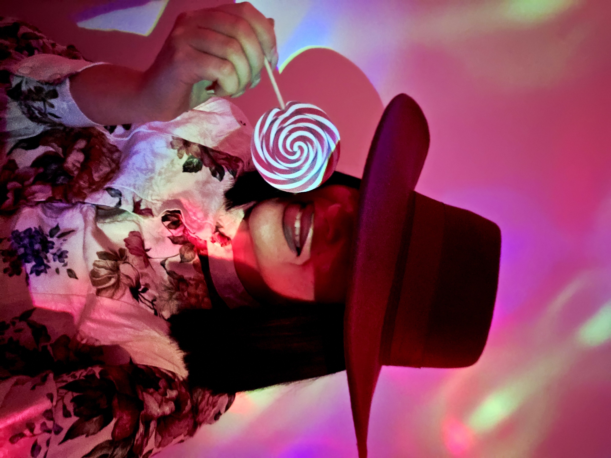 A vibrant, artistic close-up features a person, likely a woman, in an artificially lit setting. The subject's head, adorned with a dark red, wide-brimmed hat (possibly a fedora), is positioned to the right of the frame, facing slightly upwards. She wears a light-colored garment patterned with various shades of red, pink, brown, and green flowers.
Her hand, positioned towards the top left, holds a lollipop with a prominent red and white spiral design on a stick. The lollipop is held close to her face, directly in front of her slightly open mouth, hinting at the act of tasting or about to taste it. The lollipop appears to glow with a cool blue hue, contrasting with the warm, surrounding colors.
The entire scene is bathed in dynamic, projected lighting, creating a captivating backdrop. Dominant colors are intense pinks, purples, and magentas, with ethereal hints of green and blue light scattered across the background and subtly illuminating parts of her clothing, skin, and the hat. This colorful illumination creates a dreamy, stylized atmosphere. There are no specific location cues or visible text, suggesting an indoor studio setting or a carefully curated environment focused on visual aesthetics. The mood conveyed is playful and visually striking.