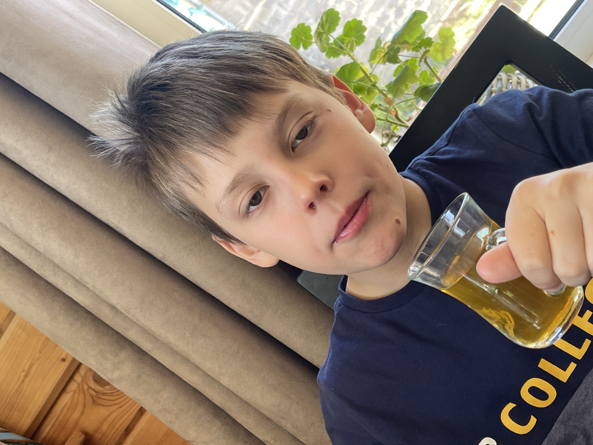 A boy holds a small glass mug of tea in front of him. He is wearing a dark blue t-shirt with yellow lettering. He has fair skin, short brown hair, and is looking at the camera. Behind him is a window with a plant visible outside. There is also a beige curtain to his left. The location is Chaikovsky, Russia.