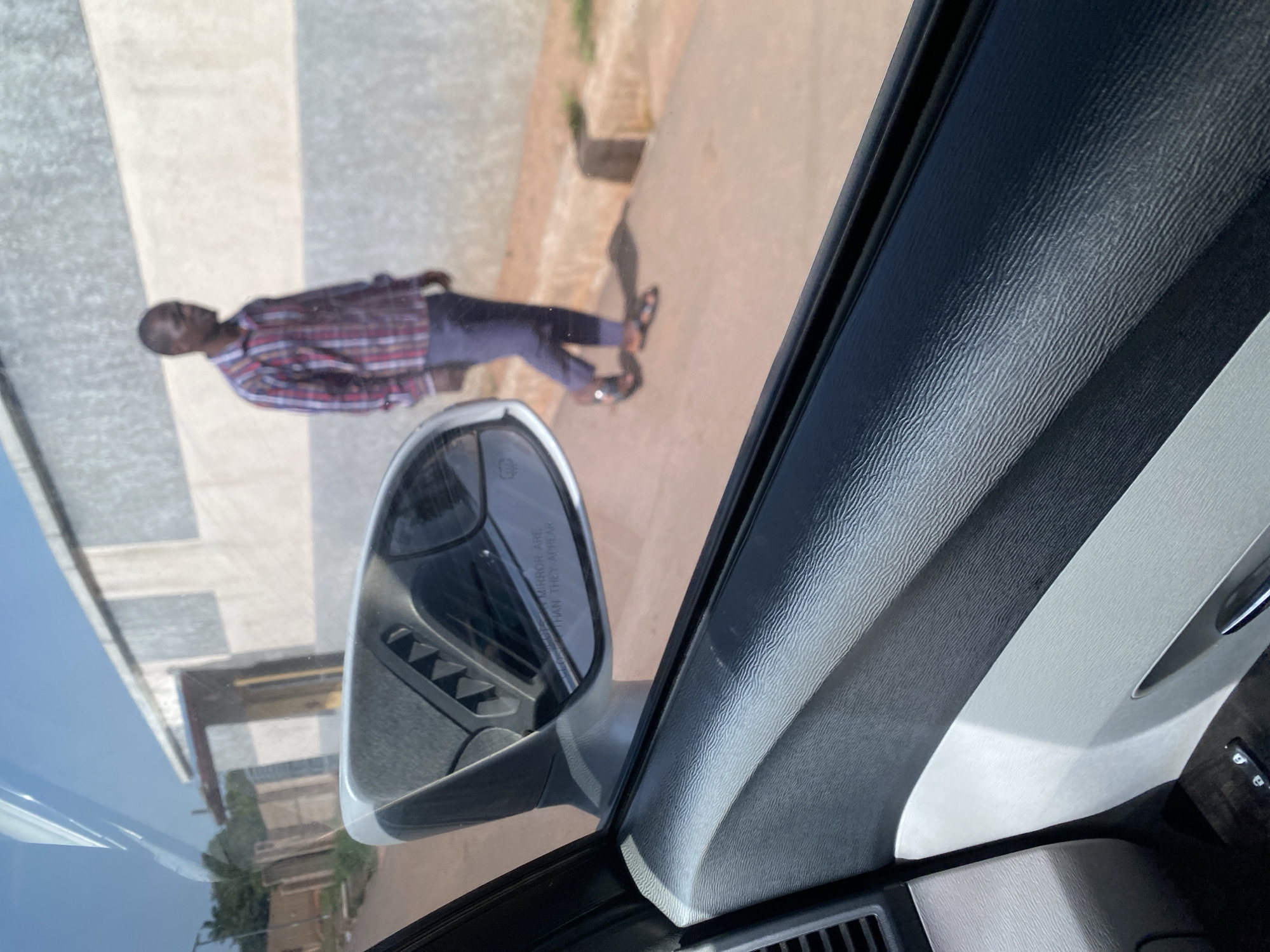 A man walks on a dirt road in front of a building with a gray and white striped facade in Bar Jirgi Summa, Nigeria. The man, wearing a red and blue checkered shirt and dark pants, is seen from the chest down. A side mirror of a car, reflecting part of the building and a dark interior, is in the foreground. The image is taken from inside the car, with the car's interior door panel visible in the lower part of the frame. The ground is a reddish-brown dirt, with a concrete curb visible. The sky is clear and blue.