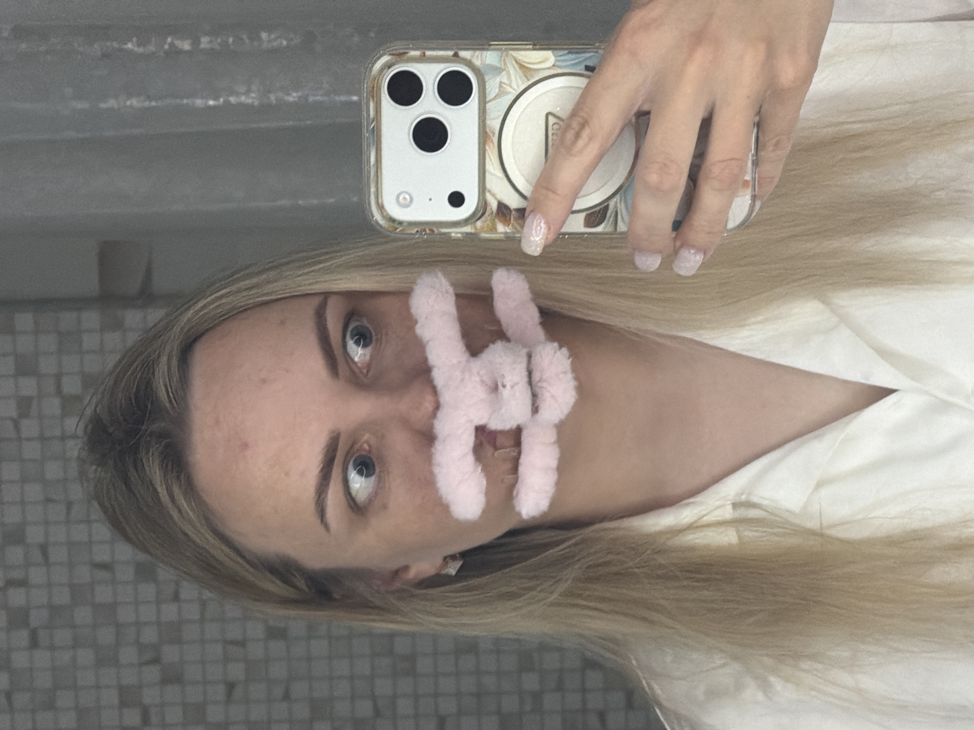 A person, appearing female, is captured in a mirror selfie, holding a smartphone with her right hand. The image is rotated 90 degrees clockwise. She has long, straight blonde hair and light-colored eyes, looking directly at the camera's reflection. Her fair skin shows a small blemish on her forehead. A whimsical pink, fuzzy, rabbit-ear-shaped object is attached to her face, covering her philtrum and upper lip area. She is wearing a light-colored, possibly white or off-white, collared shirt.
The smartphone, an iPhone model with three camera lenses and a flash/sensor, is housed in a clear case adorned with a muted floral or leafy pattern in shades of beige, brown, and light green. A circular white grip accessory is affixed to the back of the case, with the text "PopS" partially visible. The person's fingernails are painted with a light, glittery polish.
The background behind the person is a tiled wall featuring small, light grey square tiles, suggesting an indoor setting such as a bathroom. Above the reflected person's head, a plain, light-colored surface is visible. The scene takes place in Votkinsk, Russia.