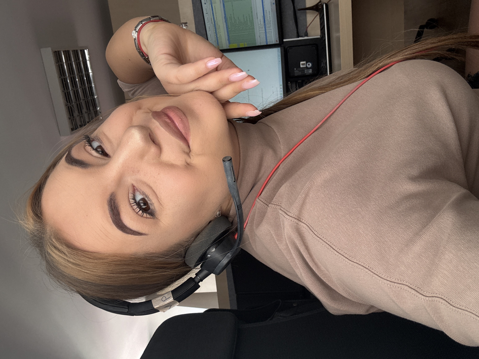 A woman, approximately 20-35 years old, with long brown hair and highlights, is depicted in an indoor office setting in Sofia, Bulgaria. She is seated in a dark office chair, wearing a beige or light brown long-sleeved turtleneck top with visible stitching. A black headset with a microphone boom is worn over her head, covering her left ear, with a red cable running down the front of her torso. She looks directly at the camera with a slight smile, resting her right hand, which has light pink manicured nails, against her chin. On her left wrist, a silver-toned bracelet with a clear stone and a red string bracelet are visible. In the background, two computer monitors display text and data, situated on a light-colored desk. Below the monitors, a black electronic device is present. A small decorative item, resembling a miniature Eiffel Tower, is partially visible on the desk to the far right. A rectangular ceiling-mounted ventilation grille is visible in the upper right background.