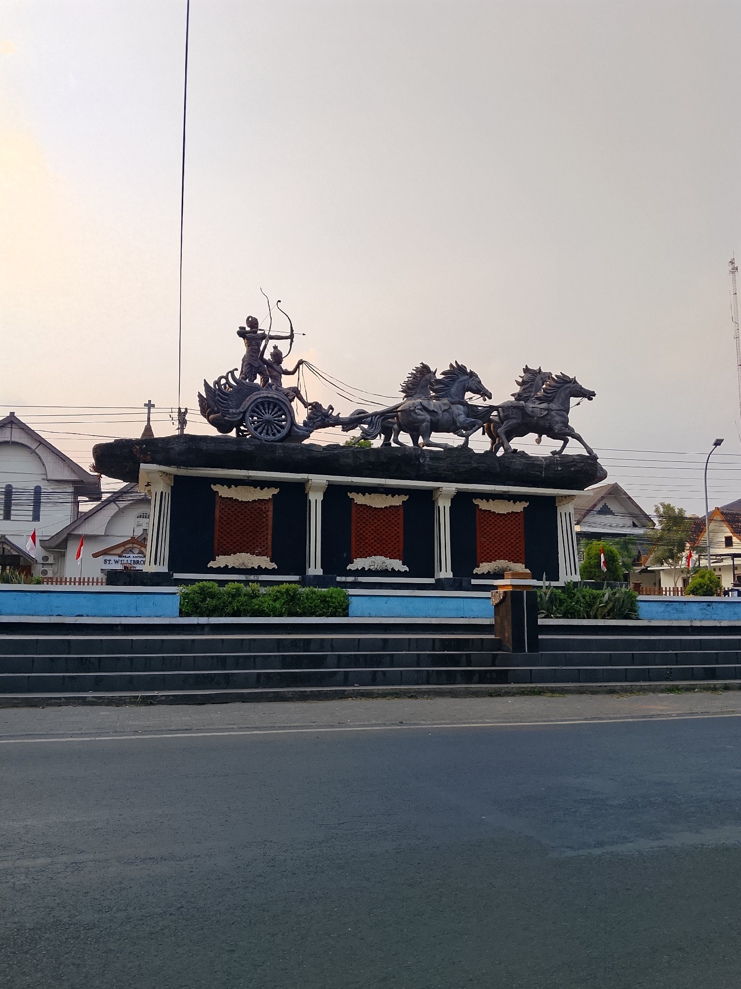 This is a medium-angle, eye-level, full-shot photograph of a monument in Balun, Indonesia. The monument depicts a chariot pulled by four horses. Two figures, one with a bow and arrow, are in the chariot. The monument is dark gray, and the horses appear to be made of metal. The monument is situated on a tiered platform with dark gray steps. Behind the monument, there are buildings, including a white building with a cross on top that appears to be a church. The sky is a light orange and gray, suggesting it is either sunrise or sunset. The foreground shows a gray asphalt road.