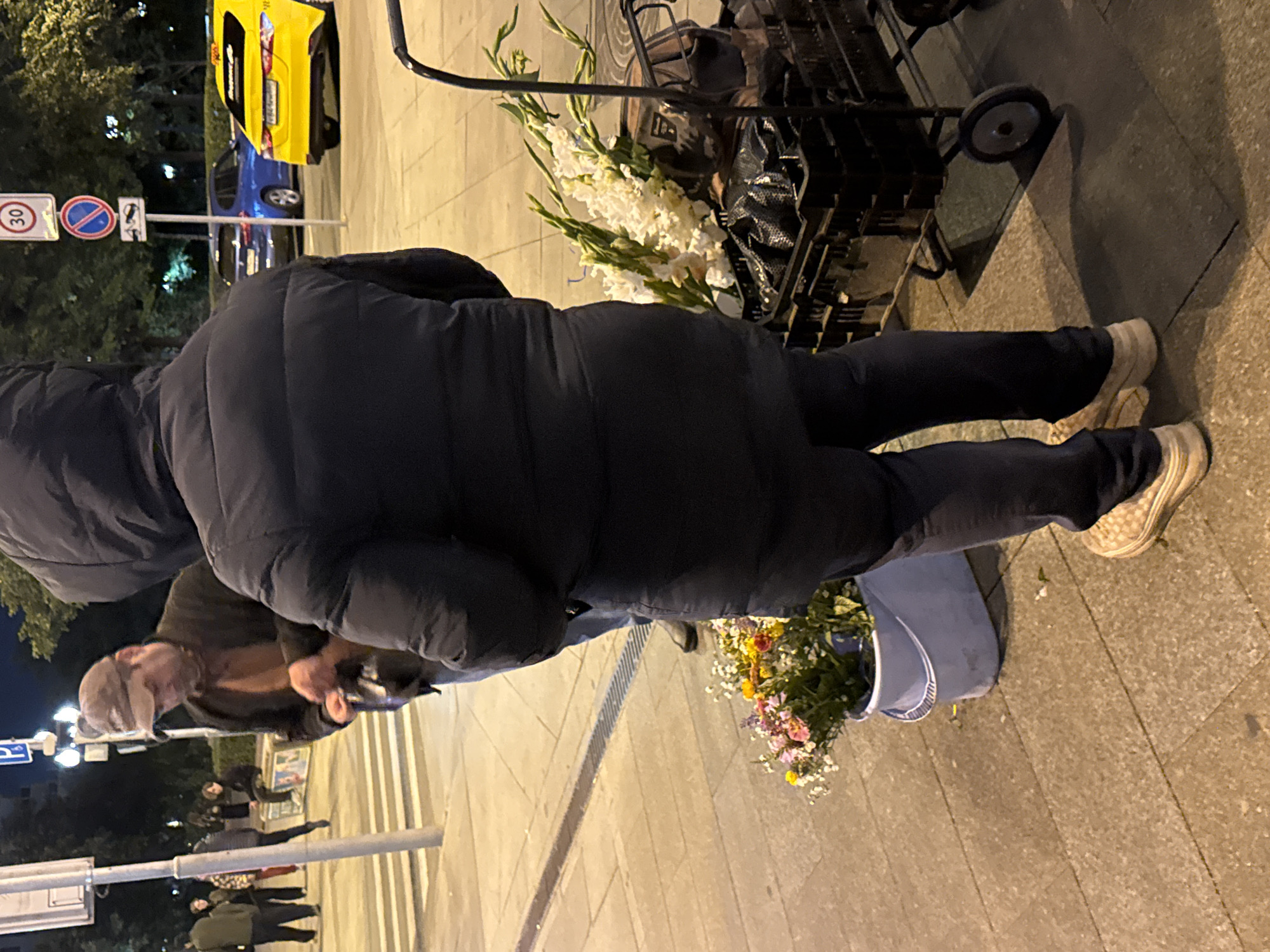 A man in a dark hooded jacket and dark pants stands in the foreground facing away from the camera. He is wearing light-colored sneakers. To his left, a bucket of colorful flowers sits on the sidewalk. Behind him, a cart filled with large white flowers and black bags is visible. In the background, a man wearing a baseball cap and a brown jacket is interacting with another person, and several other individuals are standing on the sidewalk. Behind them, streetlights illuminate a crosswalk, and cars, including a yellow taxi, are present on the road. Traffic signs are visible on a pole. The scene appears to be a street in Moscow, Russia, likely at dusk or night given the lighting conditions.
