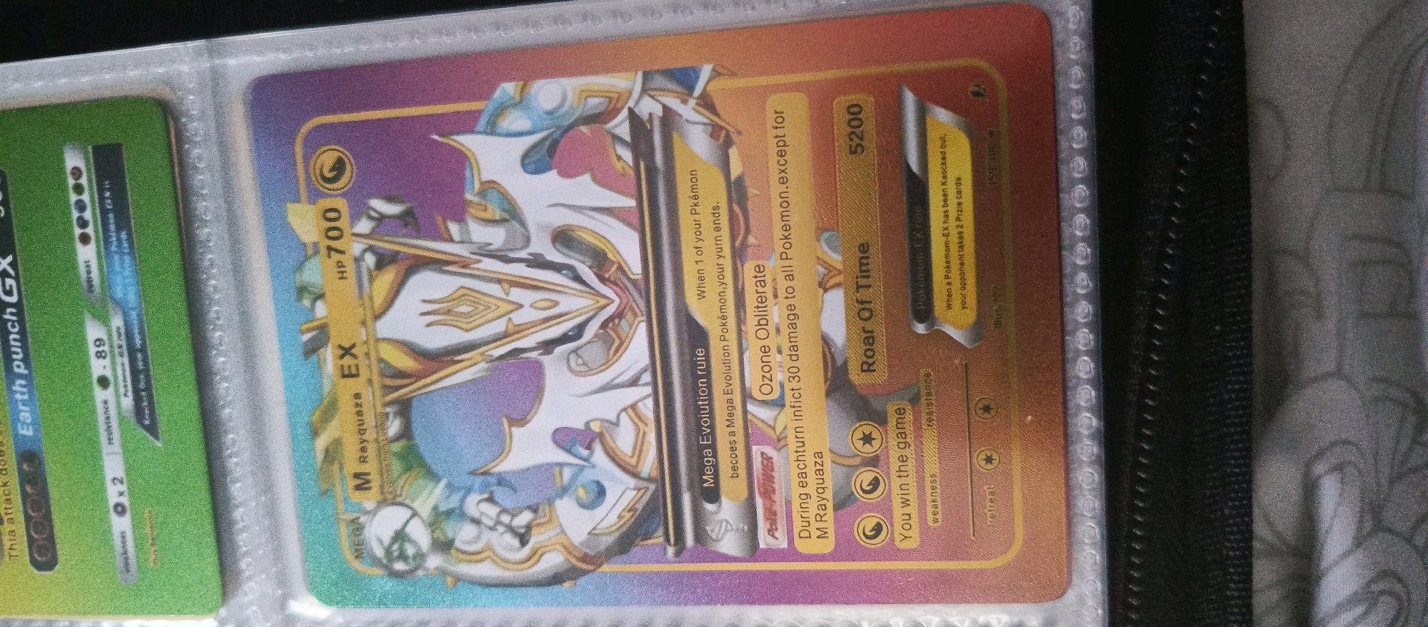 The image is a close-up, angled photograph of a Pokémon trading card. The card features a character that appears to be M Rayquaza EX, with a powerful, armored design. The background of the card transitions from purple to orange, with the character itself predominantly white and gold, accented with blue and purple. Text elements on the card include "HP700", "M Rayquaza EX", "MEGA", "Mega Evolution ruie", "Ozone Obliterate", "During each turn inflict 30 damage to all Pokemon.", "When 1 of your Pokemon becomes a Mega Evolution Pokemon, your turn ends.", "Roar Of Time 5200", "Pokemon EX Rule", "When a Pokemon-EX has been Knocked out, your opponent takes 2 Prize cards.", "You win the game", and "retreat". The card is held within a clear plastic page protector, and the edge of another card, green with white text including "Earth punch GX", is visible on the left. The background appears to be a dark binder or album. The photograph does not contain any elements that can be used to determine its location in Brwinów, Poland.