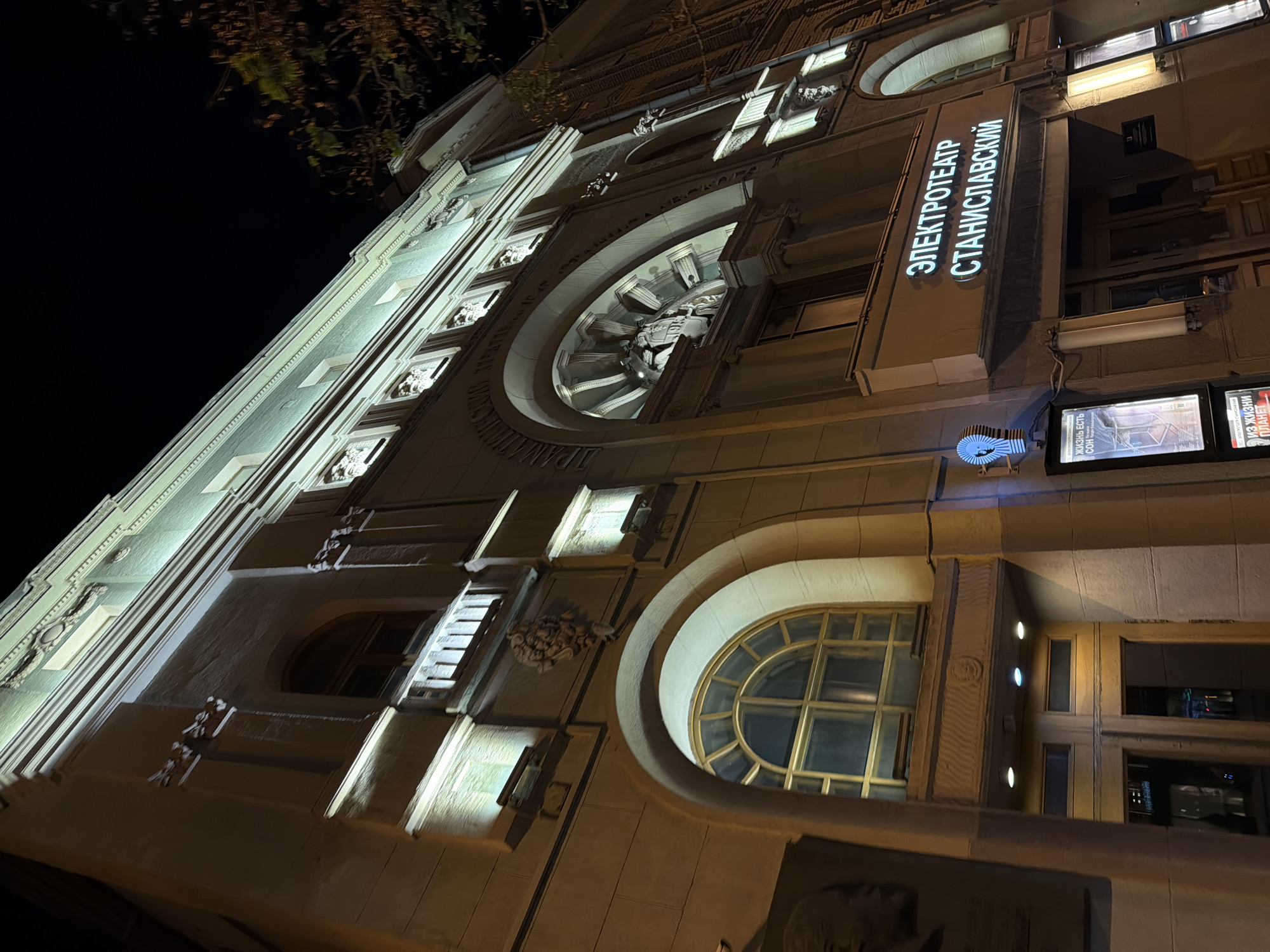 The image shows the facade of the Stanislavsky Electrotheatre in Moscow, Russia at night. The building is illuminated, highlighting its architectural details, including arched windows, decorative moldings, and signage. The theater's name is displayed vertically in neon Cyrillic script. There are posters or displays visible near the entrance. The image is taken from a low angle, looking upwards at the building.