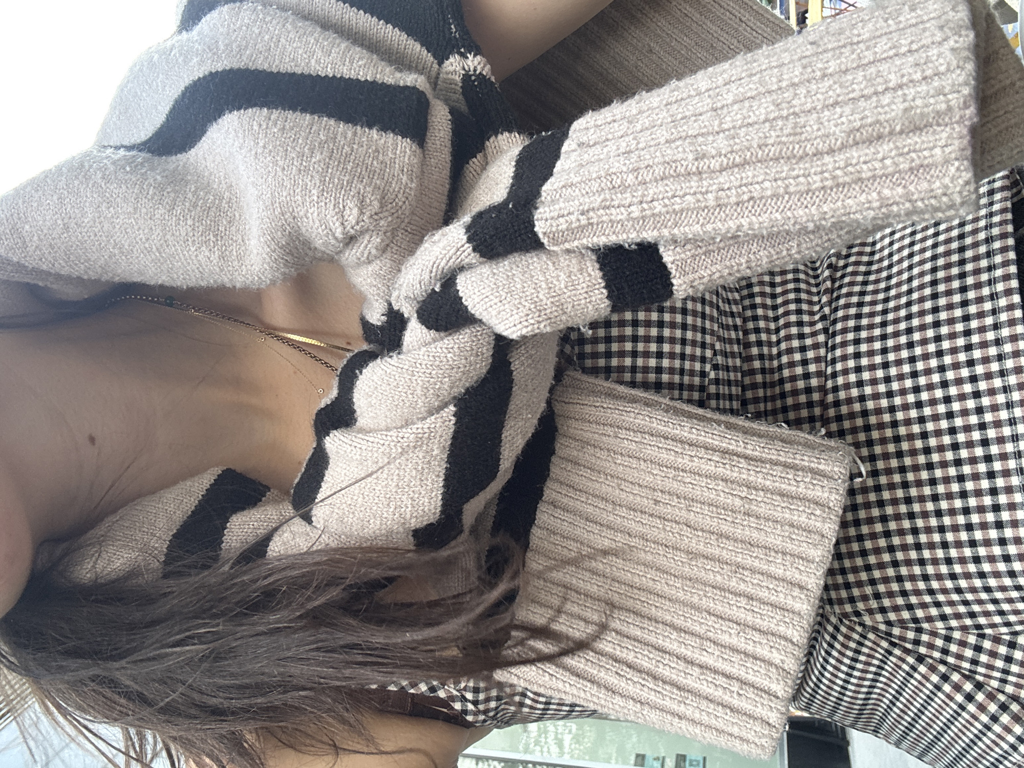 The image shows a close-up of a person wearing a checkered dress and a striped sweater draped around their shoulders. The person has long, dark hair and is wearing a delicate necklace. The background is slightly blurred, but appears to be an outdoor urban environment, possibly a street or a cafe in New York City. The lighting suggests it is daytime.