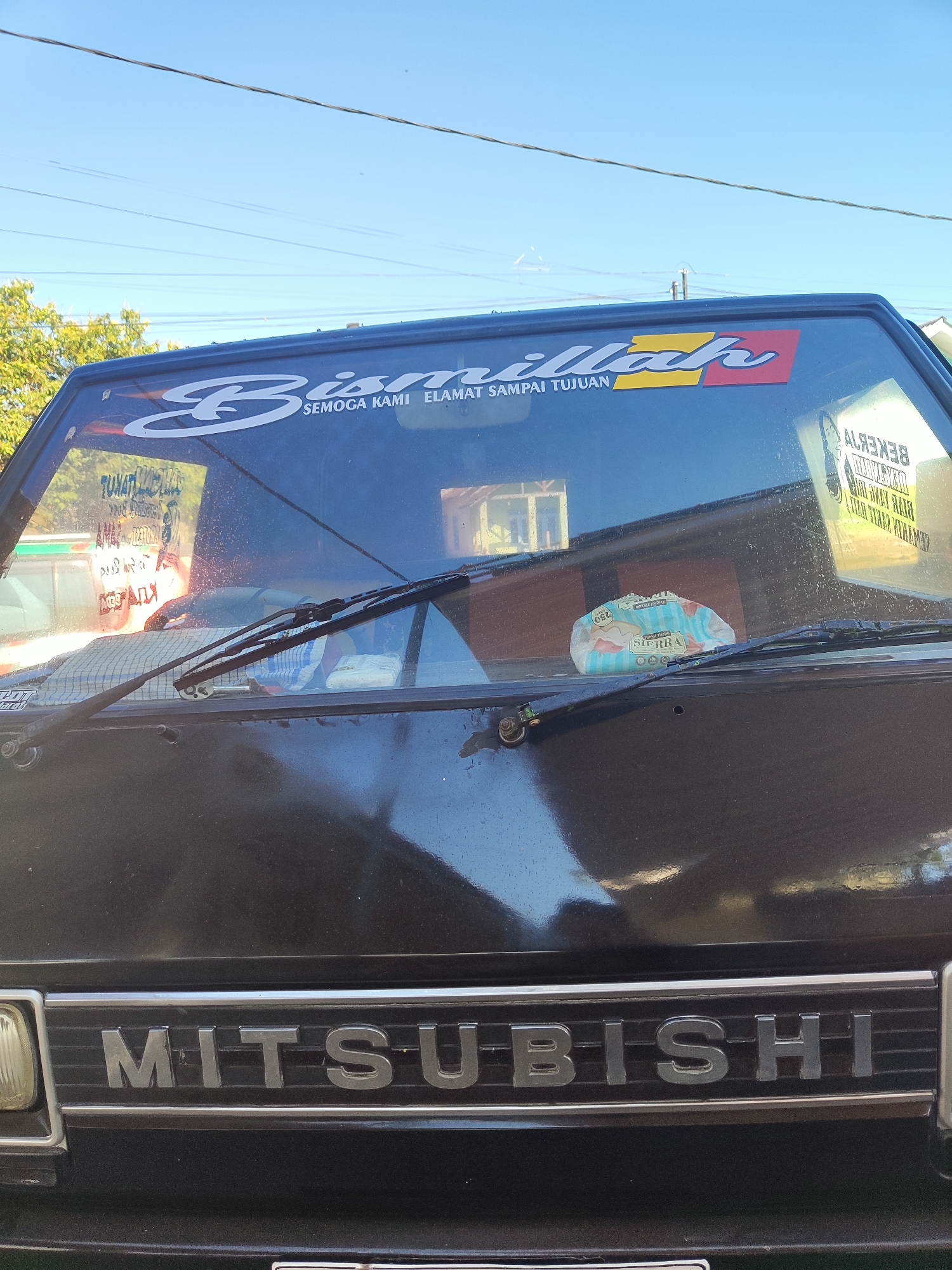 The image shows the front of a black Mitsubishi pickup truck parked outdoors under a clear blue sky, with visible power lines overhead and some trees and buildings in the background. The truck's windshield features a white decal with Arabic calligraphy that reads "Bismillah" in large script, followed by smaller text in Indonesian, "SEMOGA KAMI SELAMAT SAMPAI TUJUAN" (May we arrive safely at our destination). Below this, another sticker is visible on the windshield with Indonesian text. On the dashboard, a packet of "SIERRA" brand tissues is placed. The truck's grill prominently displays the "MITSUBISHI" logo in silver lettering. The location is identified as Pujerbaru, Indonesia.