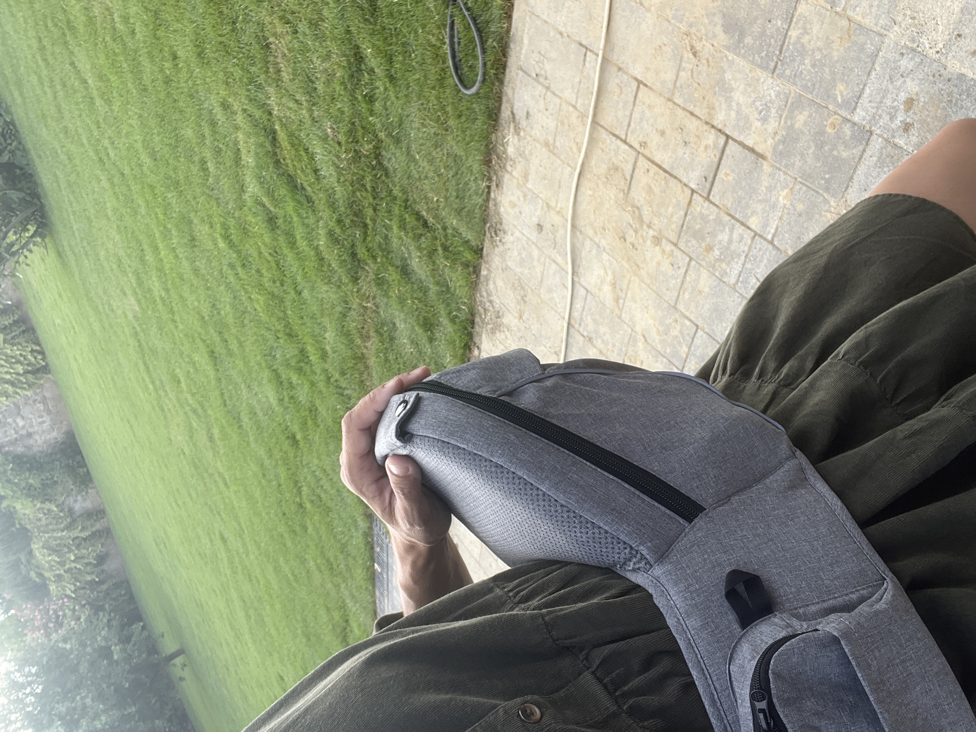 An individual wearing an olive green dress is seated outdoors, with a grey sling bag resting on their lap. The person's right hand is on top of the bag. The background consists of a sloped green lawn with trees and a stone wall at the top. The foreground is a stone patio with a white hose and a black coiled hose on the grass. The image appears to be taken in Onokivtsi, Ukraine.