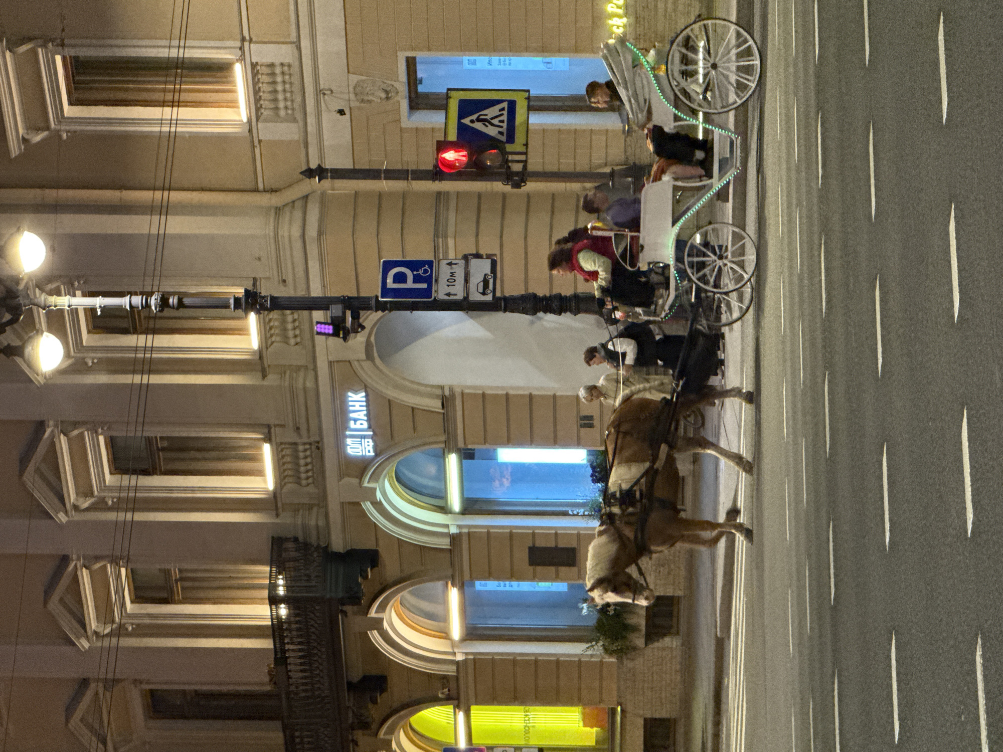 This is a nighttime street scene in Saint Petersburg, Russia. The image captures a horse-drawn carriage carrying passengers moving across a crosswalk on an asphalt road marked with white dashed lines. The carriage is white and decorated with green lights. A brown horse pulls the carriage.
In the background, a building with classical architecture is illuminated. It features large arched windows, some with yellow and blue lighting emanating from within. Above the windows, ornate architectural details are visible, and a balcony with intricate railings is present on the upper level. A street lamp with two glowing globes stands near the sidewalk.
Several individuals are visible: passengers in the carriage, a driver, and pedestrians on the sidewalk. Traffic signals, including a red light for vehicles and pedestrian crossing signs, are also present, indicating an intersection. A sign with the Cyrillic word "БАНК" (BANK) is displayed on the building, suggesting the presence of a financial institution. The overall impression is a lively evening scene in an urban environment.