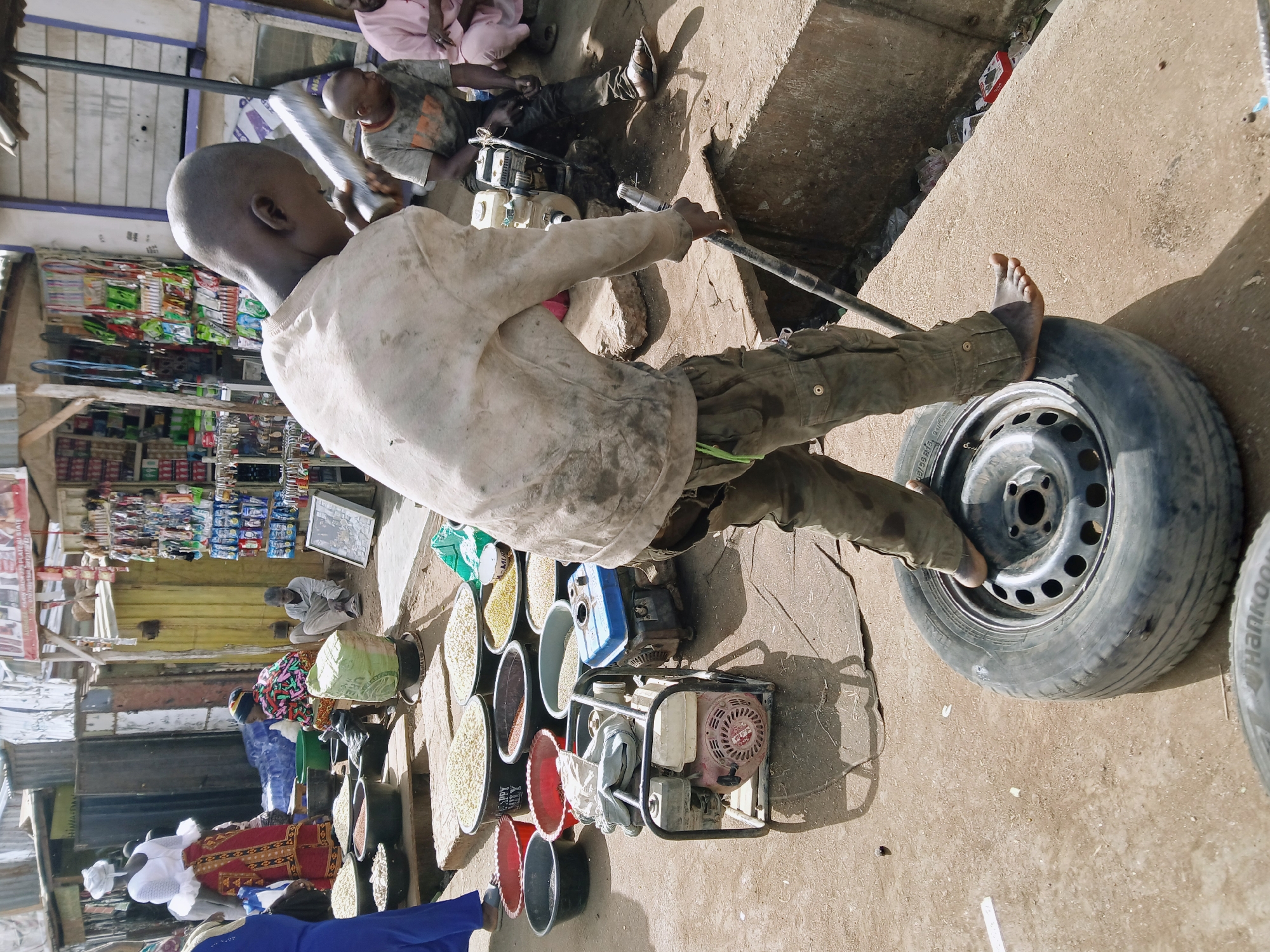 A person is standing with one foot on a car tire, holding a metal bar. Buckets of grain are visible to the left, along with a generator. Other people are visible in the background, with a shop lined with goods. The location is Rafin Par, Nigeria.