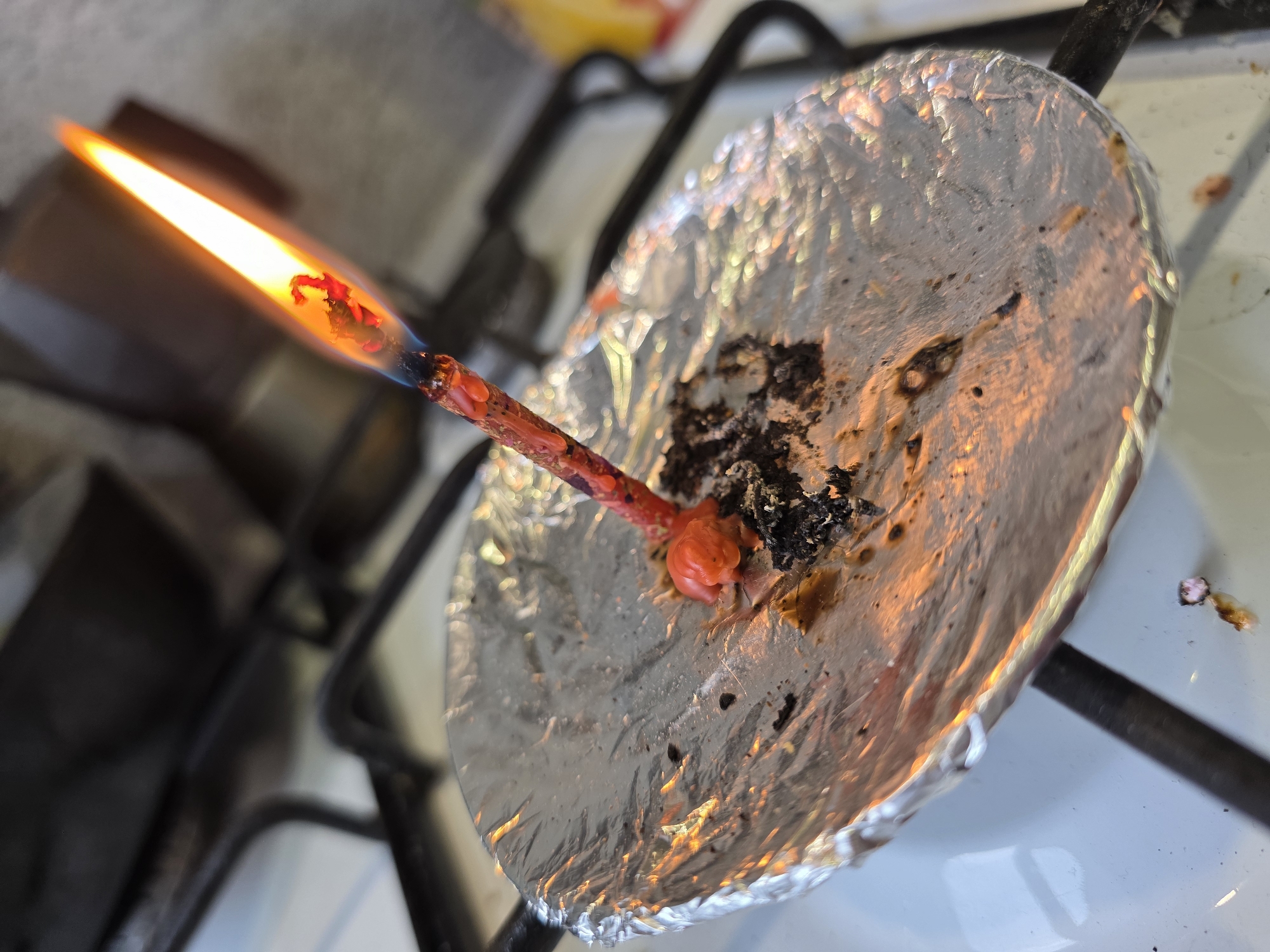 A lit candle is on top of a sheet of aluminum foil. The foil is slightly charred and appears to be on a gas stovetop burner in Chaikovsky, Russia.