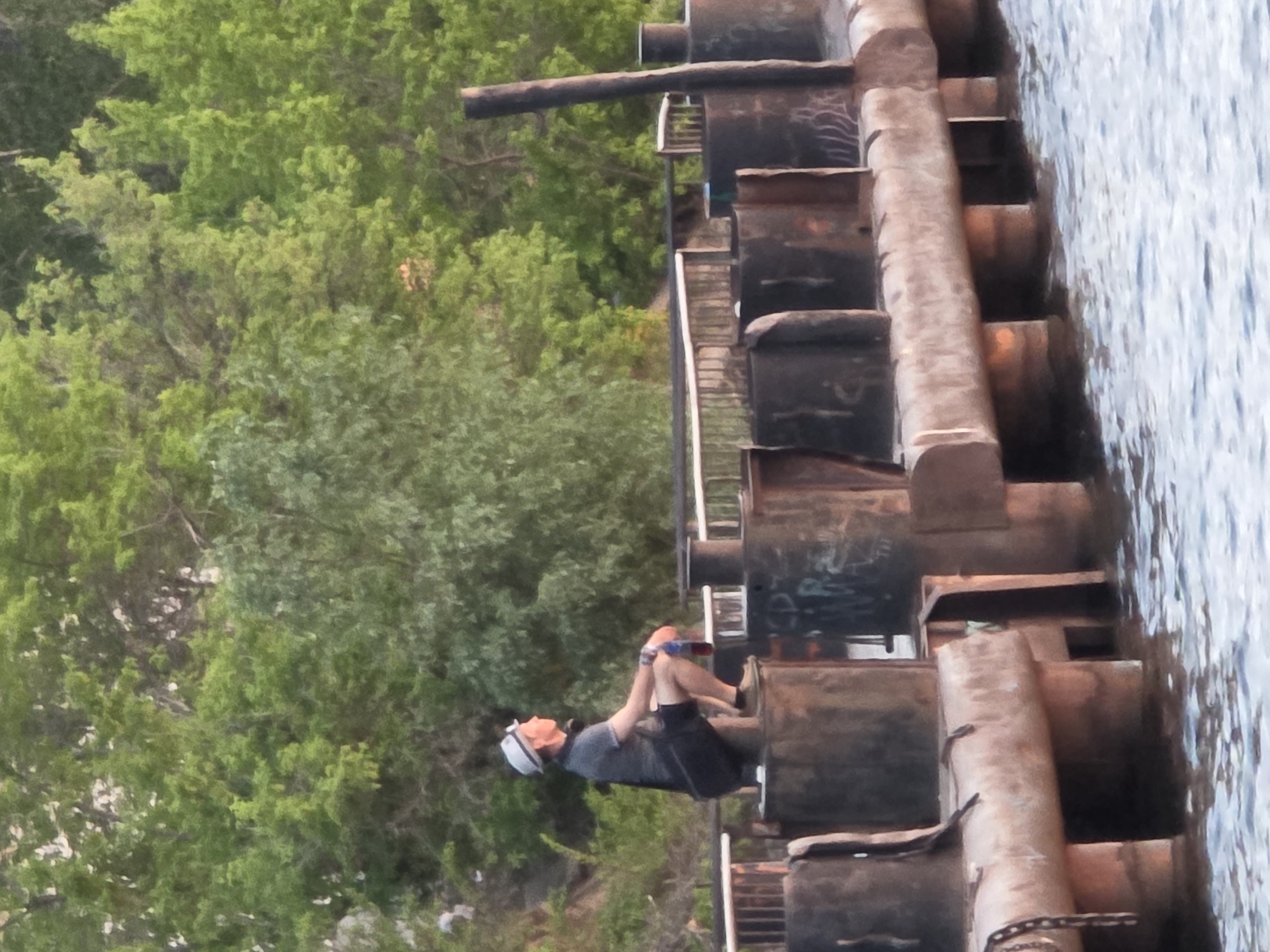 A man sits on the edge of what appears to be a section of a metal pier or dock, with green foliage to his left and water to his right. He is wearing a light-colored cap, a grey t-shirt, and dark shorts. The pier is constructed of multiple metal cylinders stacked vertically. The location is Chaikovsky, Russia.