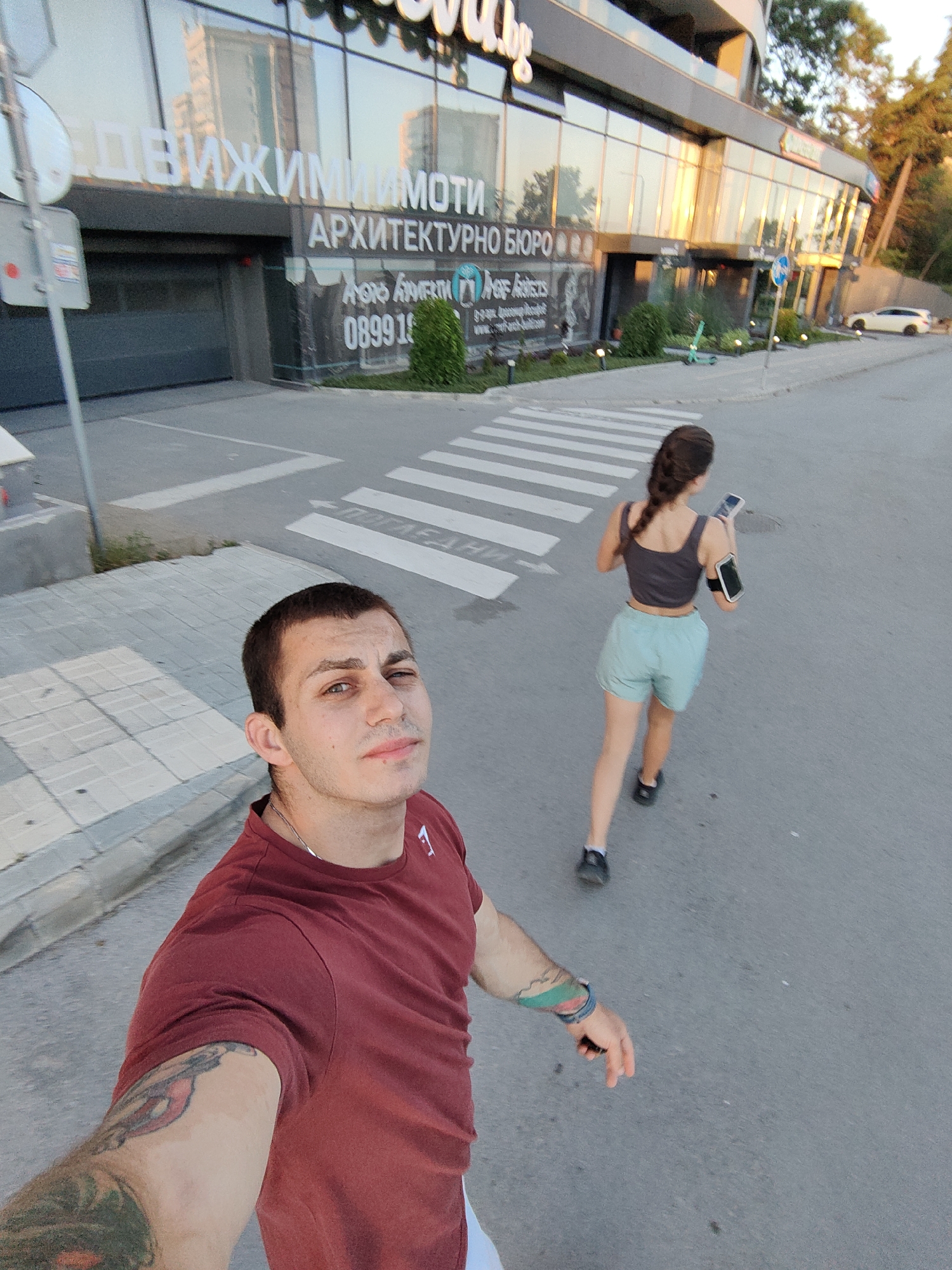 The image captures a street scene in Varna, Bulgaria, likely in the area of the newly constructed buildings visible in the background. In the foreground, a young man in a maroon t-shirt takes a selfie. He has tattoos on his left arm. Behind him, a young woman with a braided ponytail is jogging across a pedestrian crossing. She is wearing a dark tank top, light blue shorts, and a black armband with a phone attached. The background features a modern building with large glass windows, reflecting the surrounding environment. The street is paved, and there are visible pedestrian crossings and road markings. A scooter is parked near the building. The time of day appears to be late afternoon or early evening, with the setting sun casting warm light on the building.