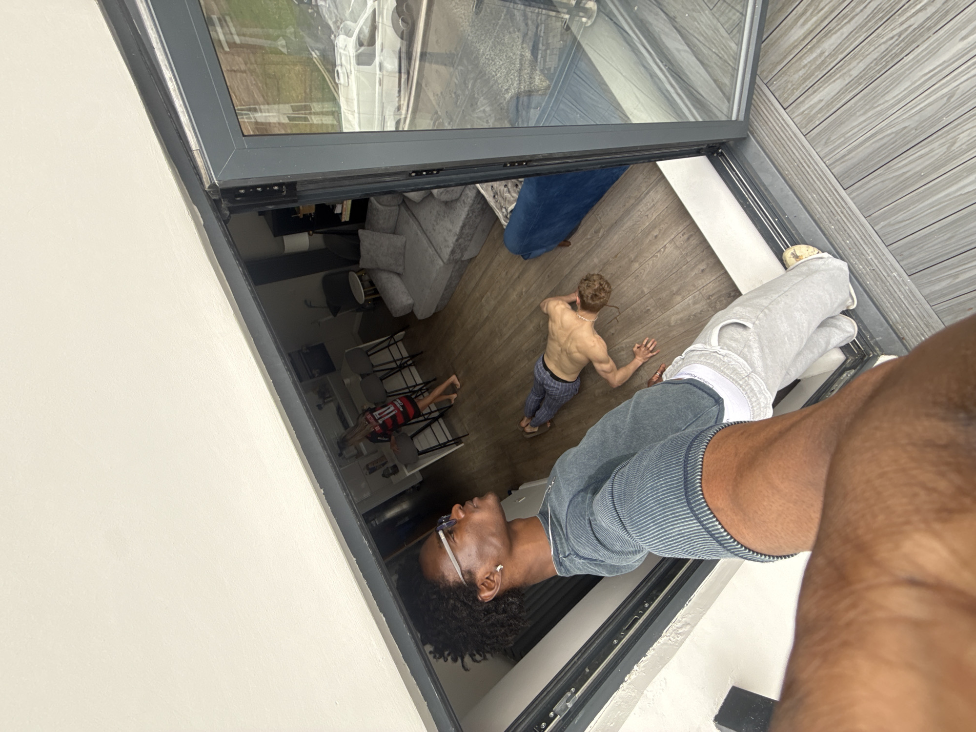 The image shows an angled view of a person holding a mobile device, presumably taking a photo or video. This person is wearing a grey and blue striped shirt, grey sweatpants with a Calvin Klein waistband, and light-colored footwear. Another person, shirtless and wearing patterned pants, is lying on the floor inside the room, with another person in a red shirt is also on the floor. The room contains furniture including a grey sofa, chairs, and a table. The photo is taken from the outside through a glass door, which reflects a view of outdoor decking and a vehicle parked in the distance. There are indications of an urban setting, potentially residential, but without further context, it is impossible to confirm that the location is London, UK.
