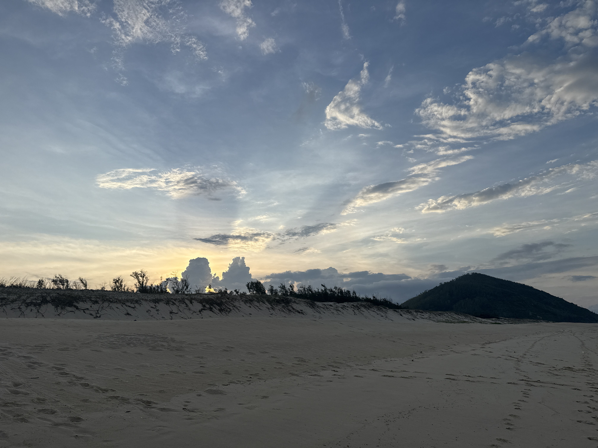This is a wide-angle, eye-level shot of a sand dune landscape under a partly cloudy sky. The foreground is a sandy beach with faint footprints and tire tracks. A low line of vegetation with sparse trees runs along the top of the dunes in the mid-ground, separating the beach from the sky. In the distance, a dark, conical mountain rises on the right side of the frame. The sky is filled with a mix of soft, wispy clouds and more defined, dark formations, with rays of sunlight peeking through in the center. The overall lighting suggests either early morning or late afternoon. The location is described as Unknown, Unknown.
