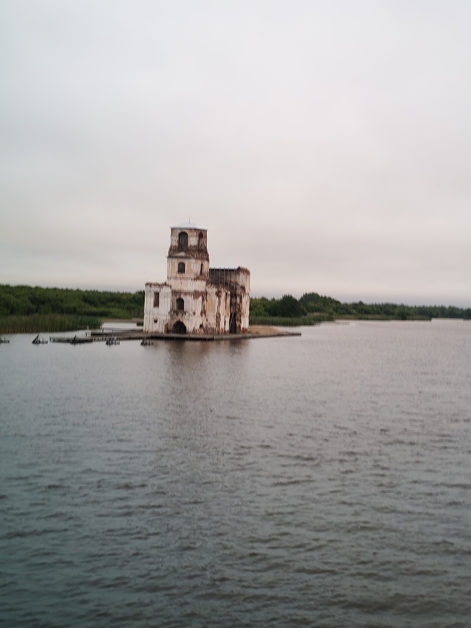 A faded, vertically oriented photograph depicts the partially submerged ruins of a white stone building, resembling a church or monastery, situated in the middle of a body of water. The building, with its tall, multi-tiered tower and arched windows, shows significant signs of decay, with crumbling walls and exposed brickwork. The water surrounding the structure is calm and appears murky, reflecting the overcast sky. A wooden pier or jetty extends from the shore towards the building. In the background, a dense line of green trees forms the shoreline, with reeds growing in the shallow water closer to the building. The overall atmosphere is one of desolation and abandonment. The image appears to be of the Church of the Virgin Nativity in Krokhino, Russia, which was flooded in the mid-20th century after the construction of a hydroelectric dam.