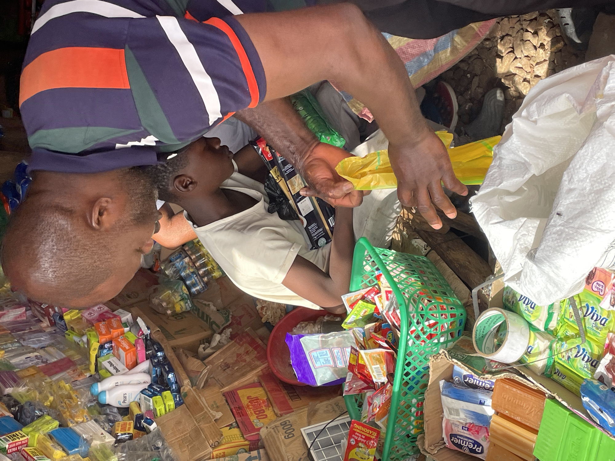 The image depicts an outdoor market scene in Akwanga, Nigeria. A man with a shaved head, wearing a striped navy blue, orange, and white polo shirt, stands adjacent to a young boy in a white shirt. The man is holding a yellow plastic bag, appearing to package goods.
In front of them is a chaotic assortment of items, including soap bars, toiletries like Pears lotion, and packaged food items. A green plastic basket is filled with smaller packets. Cartons labeled "JUMBO" and "600g" are visible. Rolls of tape and stacks of Viva Plus products are also present.
The ground is uneven and covered with loose rocks and some debris. The scene is lit by natural daylight.