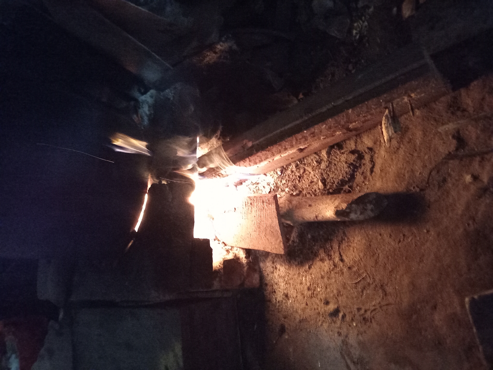 The image depicts a dimly lit, enclosed space, likely an outdoor furnace or forge in Claket, Indonesia. A fire burns brightly in the center, illuminating a wooden plank positioned over a metal trough. Debris, including dirt and small stones, covers the ground. The overall impression is one of industrial activity or a domestic cooking area, with evidence of heat and a rough, unrefined environment. The lighting suggests it is either nighttime or a heavily shaded interior.