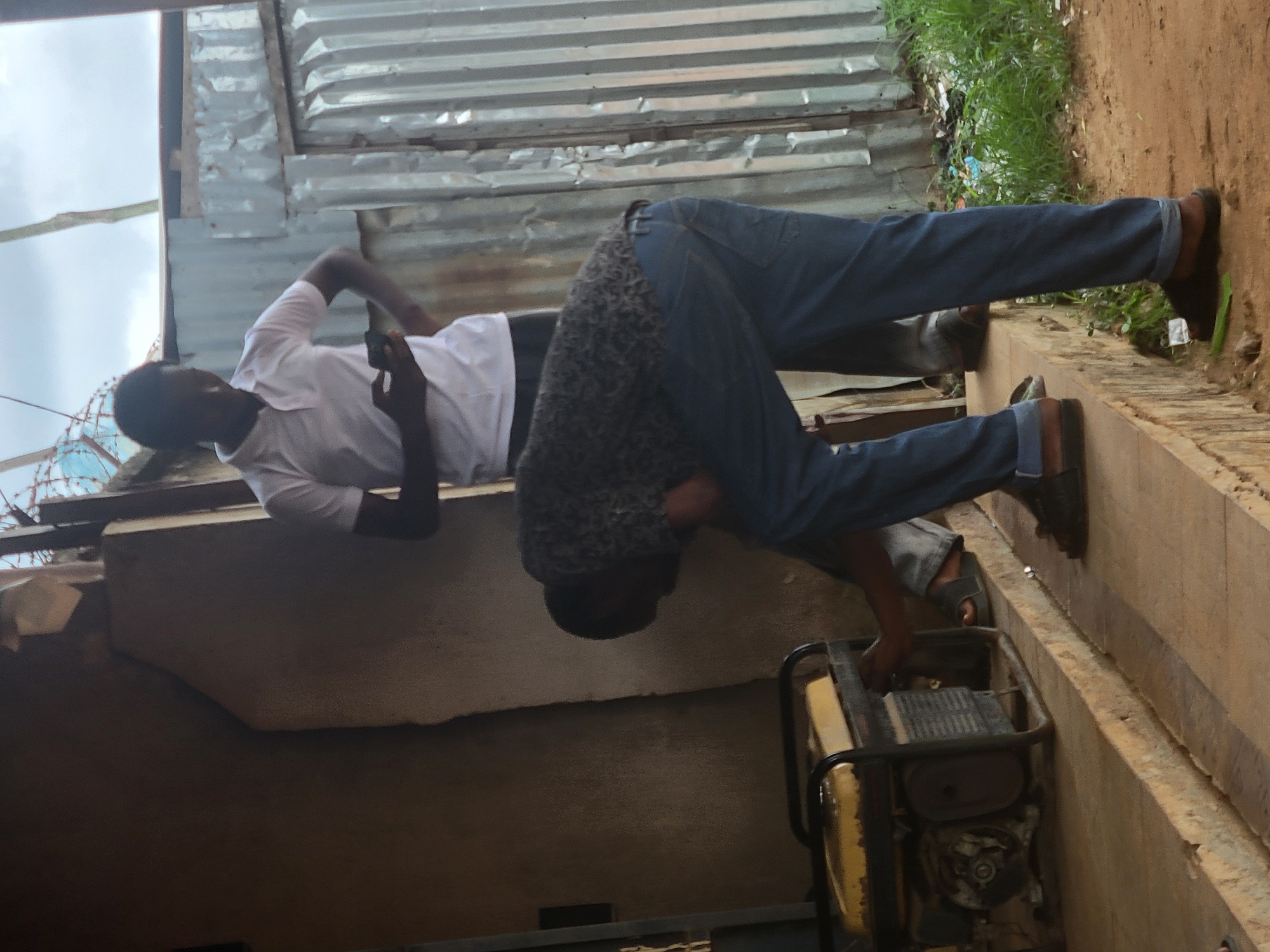 The scene depicts two men outdoors in Keffi, Nigeria. A dark-skinned man in a white polo shirt and dark trousers stands on a concrete platform, holding a smartphone in his left hand with his right hand on his hip. Another dark-skinned man, wearing a patterned shirt and blue jeans, is bent over and actively engaging with a yellow and black portable generator situated on the same concrete platform. Both men wear dark sandals. The background features a textured, light brown wall to the left, and a corrugated metal sheet wall to the right, beneath an overcast sky. Green grass and dirt are visible on the ground level below the platform.