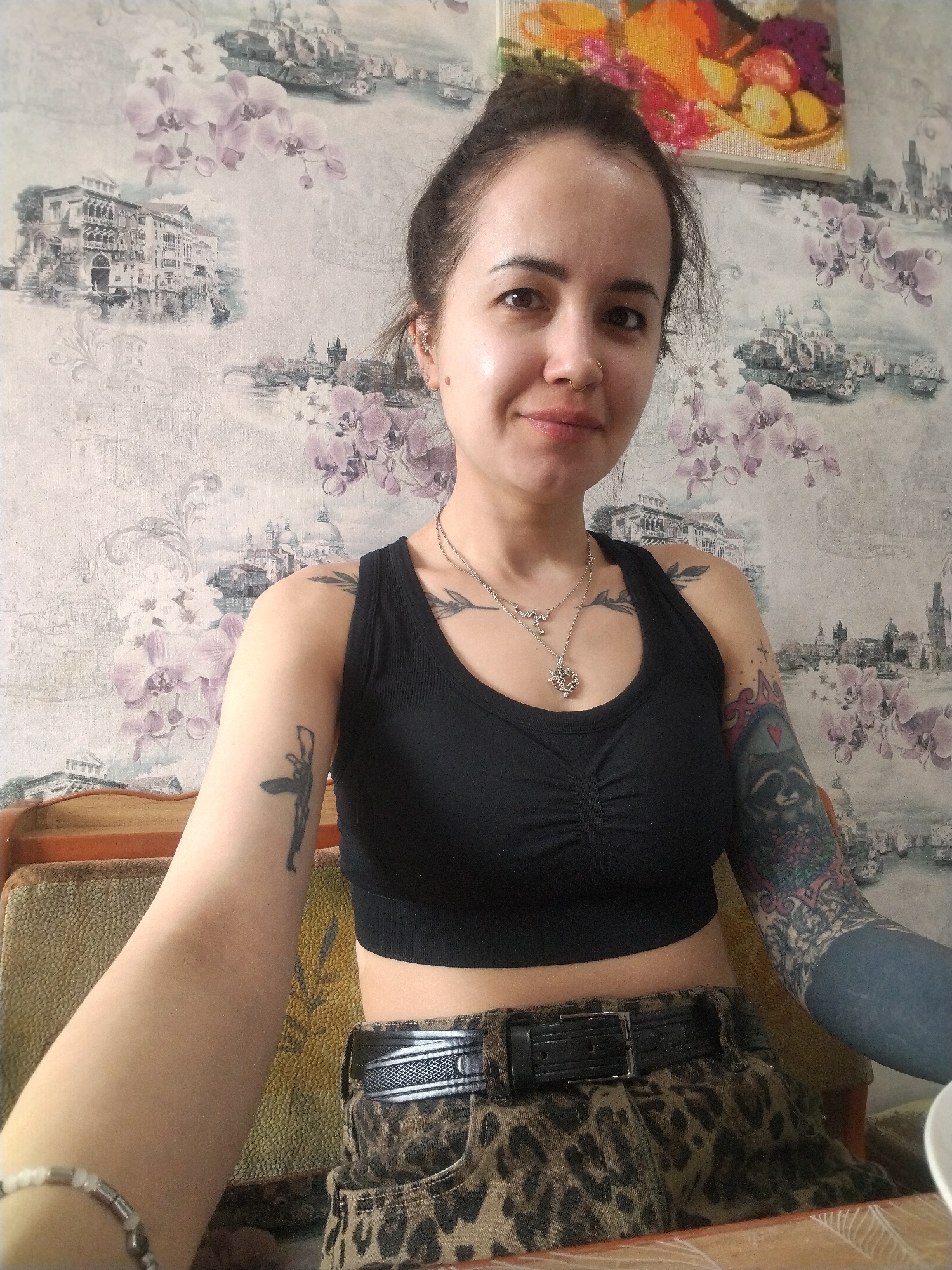 A female individual is depicted in an indoor setting, presenting a subtle smile directly towards the camera. She has dark hair styled upwards, a gold-colored hoop nose ring on her right nostril, and a small mole on her right cheekbone. She is wearing a black ribbed crop top, leopard print high-waisted shorts with a black belt, two silver-colored necklaces (one with a multi-layered pendant, the other a simple chain), a silver-colored stud earring in her right ear, and a white beaded bracelet on her left wrist. Her left upper arm features a dark tattoo of an abstract figure, and her left forearm is largely covered by a solid dark blue-black tattoo sleeve. Her right arm displays a dark foliage-like tattoo on the shoulder and a vibrant, multi-colored sleeve tattoo on the forearm, including a raccoon head within a heart motif surrounded by intricate patterns. The background wall is adorned with wallpaper depicting sepia-toned illustrations of European cityscapes, notably Venice-like scenes with canals and Baroque architecture, and a cityscape with a bridge and towers resembling Prague, all interspersed with light purple orchid blossoms. A rectangular framed artwork, possibly a cross-stitch, depicting a colorful still life of fruits and a teacup, hangs on the wall above the individual's head. Portions of a textured, light brown upholstered chair or sofa are visible to her sides, and a light-colored table surface is in the immediate foreground. The illumination is consistent with ambient indoor lighting. This scene is situated within an unknown residence in the city of Unknown, Unknown.