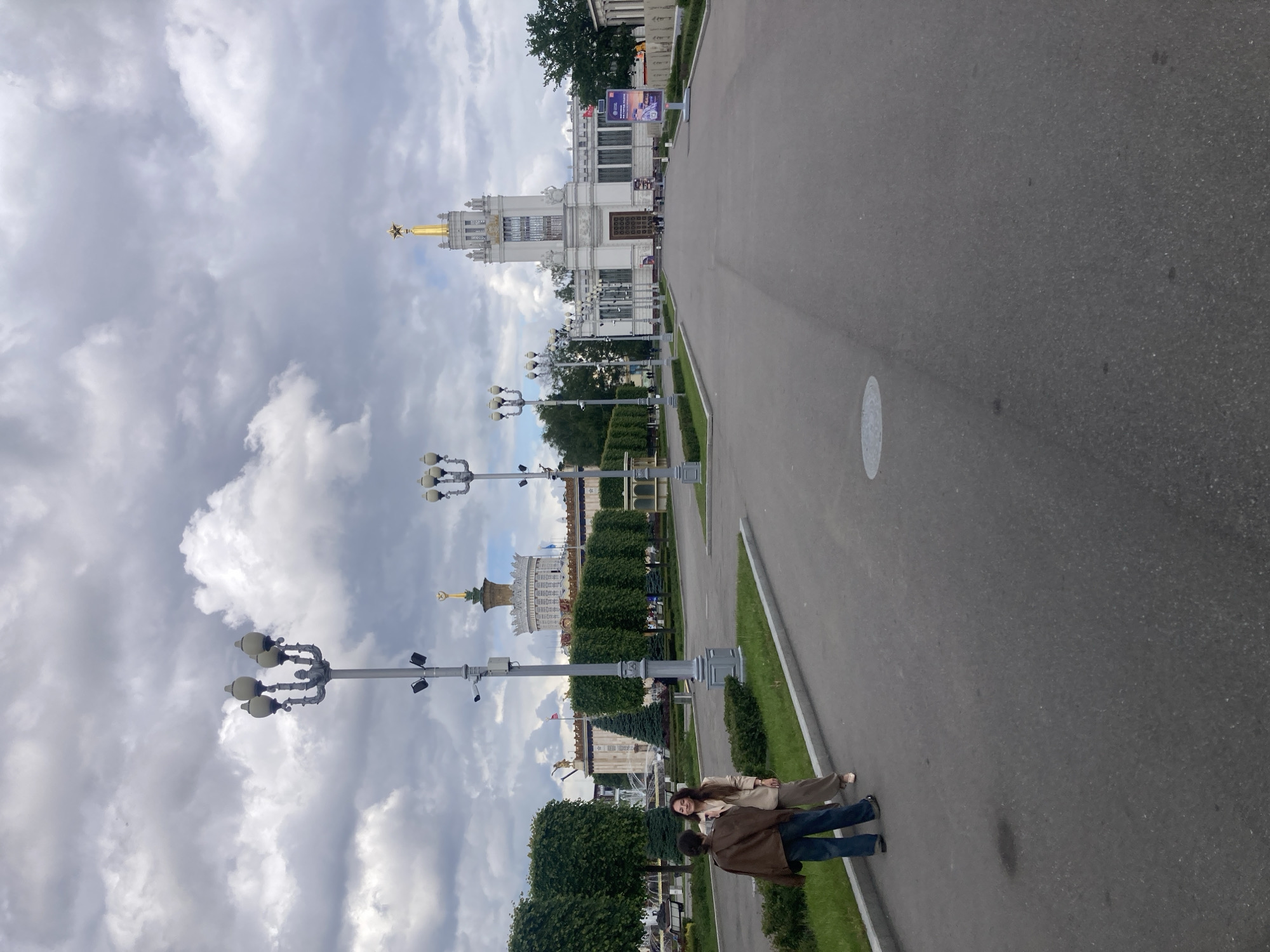 A wide, slightly downward-angled shot depicts a paved road in Moscow, Russia, flanked by meticulously manicured green spaces and elaborate lampposts. The road extends towards a large, ornate building topped with a gold-colored spire and a prominent star. The sky above is overcast, filled with dense, grey clouds. A man and a woman are walking on the left side of the road, with the woman slightly ahead. The man is wearing a brown jacket and blue jeans, while the woman is wearing a light tan jacket and matching pants. The buildings and lampposts on either side of the road are set against the cloudy sky, creating a contrast between the architectural details and the overcast weather.
