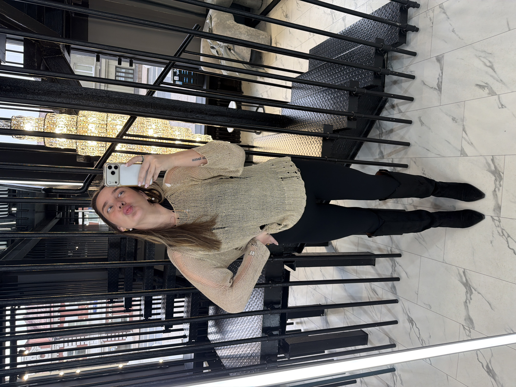 A woman takes a selfie while standing on a black metal staircase. The staircase is adjacent to a wall of windows that reflect an urban landscape. She wears a beige sweater, black pants, and black boots. A chandelier hangs above her. The flooring appears to be marble. The image may have been taken indoors in Moscow, Russia.