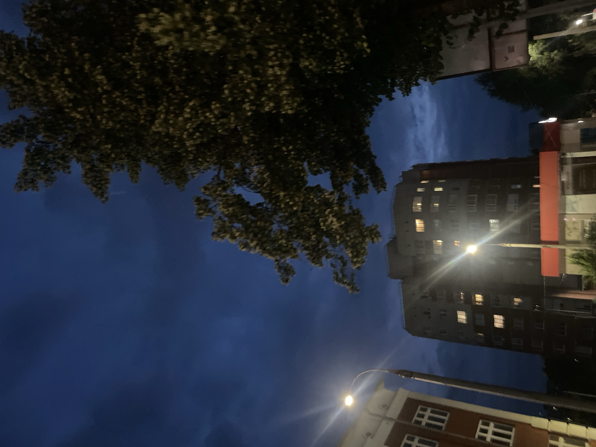 This is a low-angle, nighttime photograph taken in Votkinsk, Russia. The scene depicts an urban environment under a dark, cloudy sky. The image is dominated by the deep blue of the twilight sky, with scattered clouds creating a textured backdrop.
In the foreground, the silhouette of a large tree with dense foliage extends from the top left of the frame. Below the tree, a tall, multi-story building is visible, its windows illuminated with warm, yellow light, suggesting occupancy. To the right of this building, a portion of another building with red accents is partially visible.
A streetlamp with a bright light source is prominently featured in the lower half of the image, casting a glare across the scene and illuminating the facade of a lower building in the bottom right, which features a repeating pattern of white-framed windows.
The overall mood is one of quiet urban twilight. The time of day is clearly night, and the cloudy sky might indicate overcast weather. The presence of illuminated windows and streetlights points to a city or town setting. No people are visible, and there is no discernible text in the image.