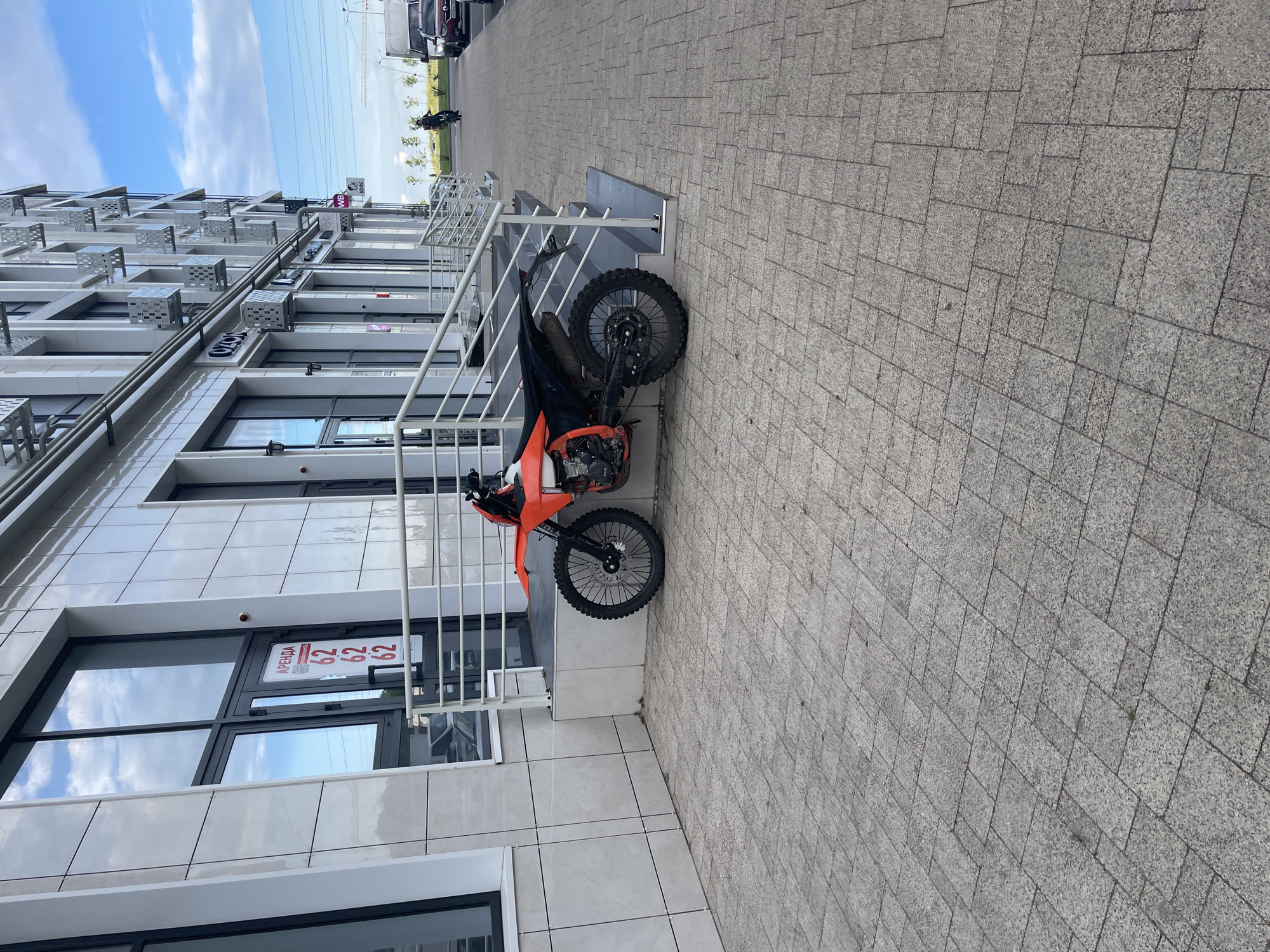 An outdoor, low-angle, eye-level shot captures a dirt bike parked on a paved area in front of a modern, multi-story building. The building's facade features white tiled panels and large glass windows. A sign on one of the windows reads "АРЕНДА" (rent) with numbers "62 62 62". A metal railing runs along the stairs leading up to the building's entrance. The ground is paved with grey square and rectangular tiles. In the background, a person on a bicycle is visible on the sidewalk, and cars are parked along the street. The sky is partly cloudy. This scene is located in Stavropol, Russia.