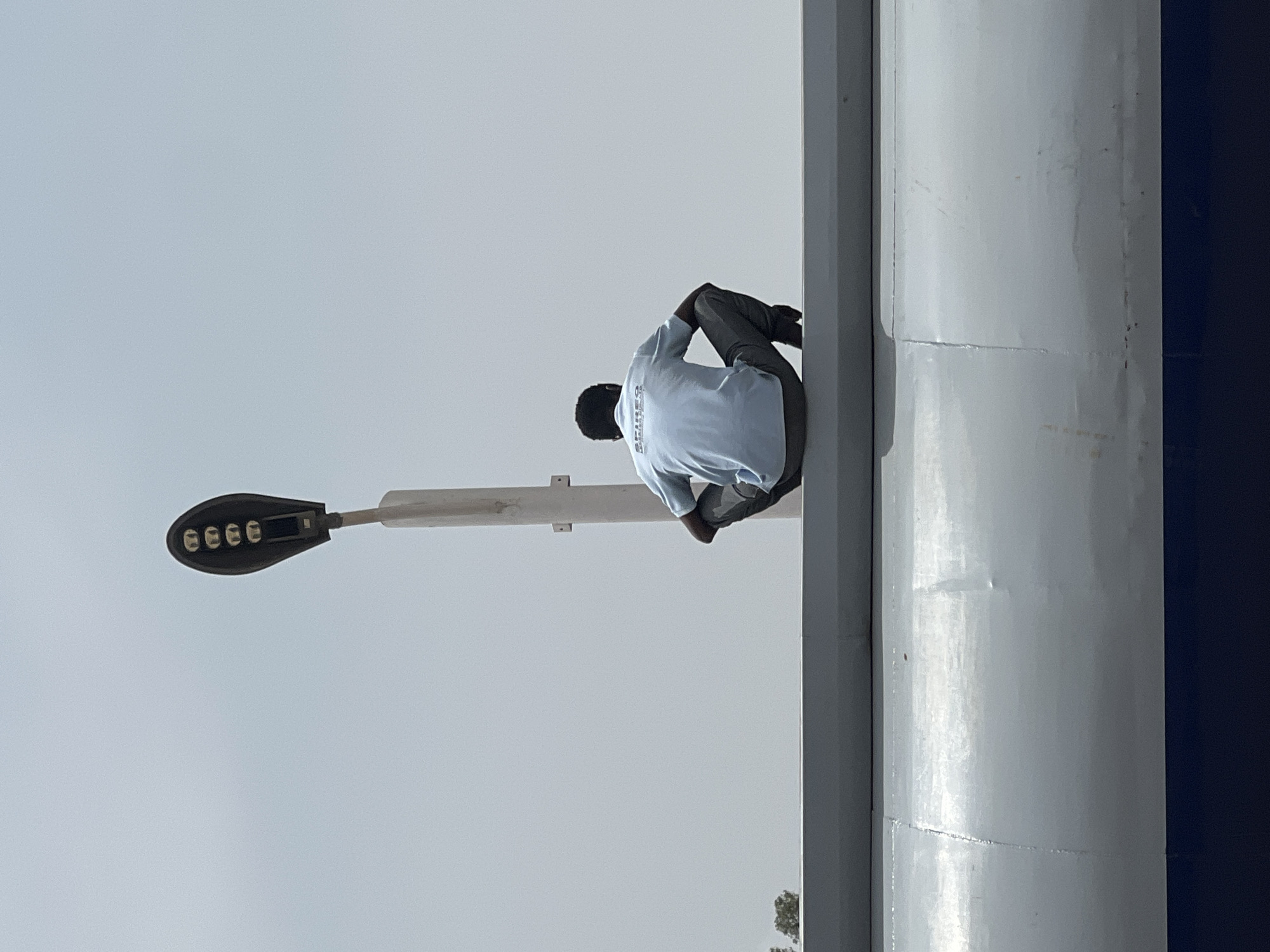 A man in a light blue shirt and dark pants is perched on a horizontal beam connected to a street light pole in Gura Top, Nigeria. The street light itself has a black head with several visible LEDs. The pole is light gray and appears to be metallic. To the right of the man and the pole, there's a large, cylindrical structure, also light gray, with a navy blue panel visible on its right side. The background is a pale sky. There is some greenery visible near the bottom of the image. The man is turned away from the camera.