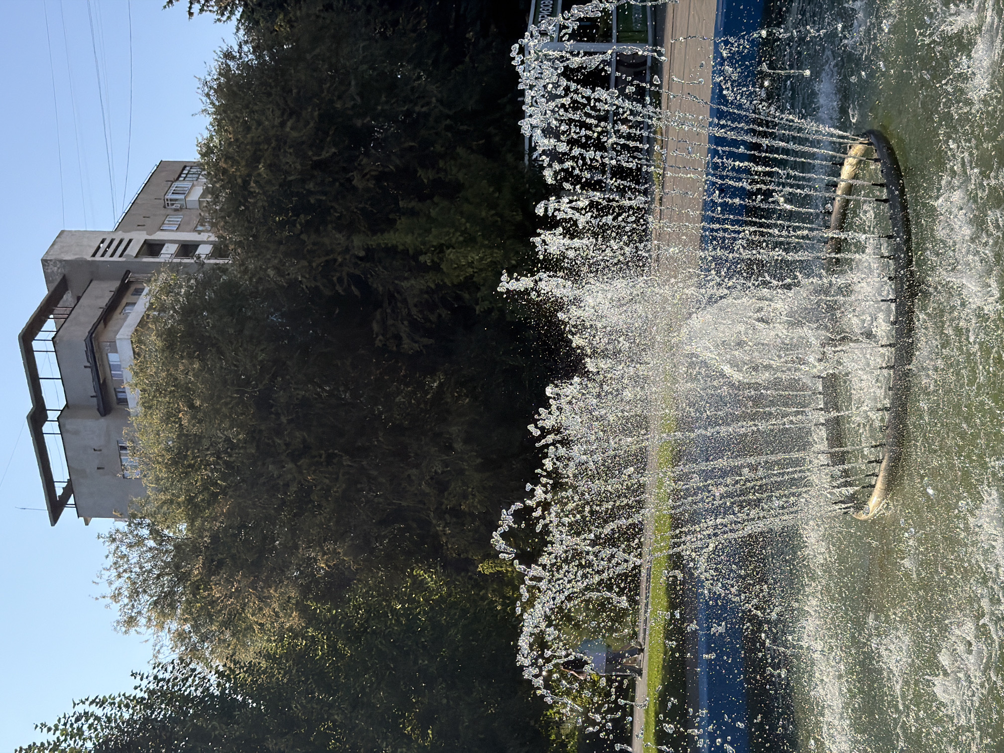 The image depicts a sunny outdoor scene, likely in a park or public square, with a fountain in the foreground. Water sprays upwards and outwards from a circular metallic structure submerged in the fountain's basin, creating a shimmering effect. In the middle ground, a paved pathway is visible, with a strip of green grass bordering it. Two individuals, their features indistinct, are walking along the pathway. Behind them, a dense line of green trees forms a backdrop. Rising above the trees is a multi-story building with a flat roof, characterized by its beige and gray facade and balconies. The sky is clear and blue. The scene is consistent with the area around the "Druzhba" fountain in Volzhsky, Russia.