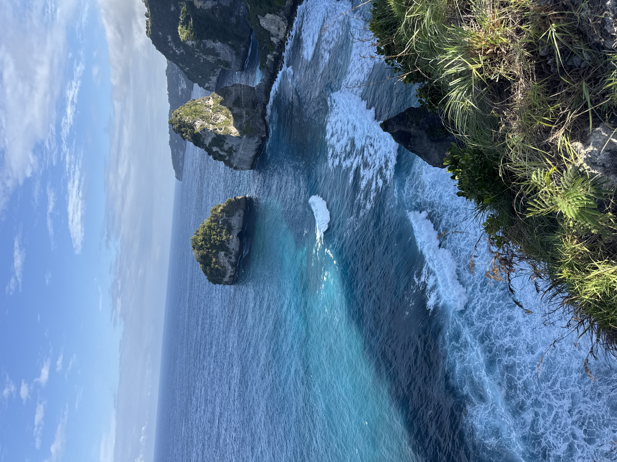 A high-angle, full shot shows a vibrant blue ocean meeting a rugged coastline on the island of Nusa Penida, Indonesia. Two large, rocky islets covered in lush green vegetation rise from the turquoise water, their sheer cliffs catching the sunlight. Waves crash against the shore, creating white foam that contrasts with the deep blue of the sea. In the foreground, a grassy cliff edge is visible, with dry grasses and green bushes reaching towards the water. The sky above is a clear blue, dotted with a few wispy white clouds. The scene conveys a sense of natural beauty and the raw power of the ocean.