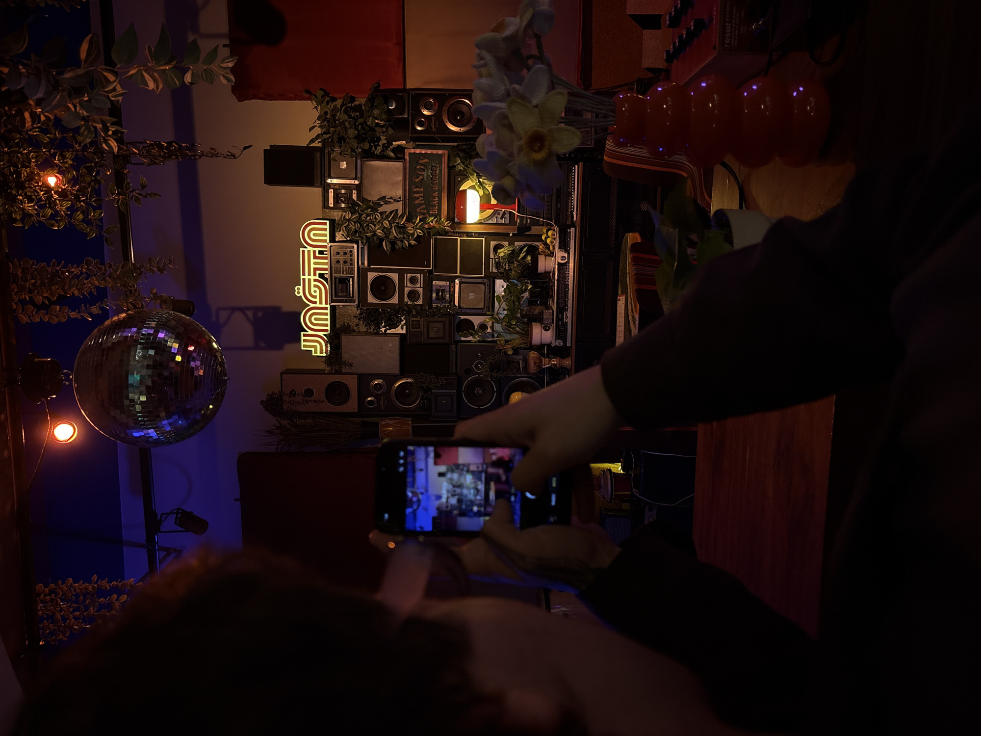 The image depicts an interior scene in Belgrade, Serbia, possibly a bar or club with eclectic decoration. A disco ball reflects light near the left, set against a blue-lit wall. The central element is a wall arrangement composed of various vintage speakers, radios, and electronic components, interspersed with plants and a neon sign reading "JUST OUT." A person is holding a smartphone, taking a picture of this arrangement. A second person is partially visible in the foreground. Red balloons are near the top right, and other elements of the bar are visible in the background. The lighting is dim, creating a moody atmosphere.