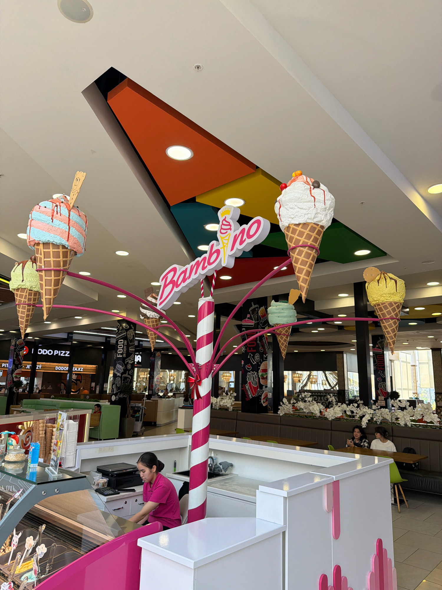 This is a medium shot, taken indoors, of a colorful ice cream parlor, likely located in the city of Pavlodar, Kazakhstan. The primary focus is a large, decorative structure resembling a stylized tree adorned with oversized, whimsical ice cream cones. These cones are made of artificial materials and are suspended from pink, curving supports. The ice cream cones are varied in color and topping. The name "Bambino" is prominently displayed in pink, stylized lettering above the ice cream cone display.
In the foreground, a young woman wearing a bright pink polo shirt is working behind an ice cream counter. She is seated and appears to be operating a cash register or calculator. The counter itself is white with pink decorative elements that mimic dripping ice cream. A display case filled with various ice cream flavors is visible to her left.
The background reveals a food court or shopping mall environment. Several restaurant storefronts are visible, including one with signage that reads "PODO PIZZ" and another with "DODOF.KZ". Tables and seating areas are present, with a few patrons visible, including two young women seated at a table in the background. The ceiling of the establishment features a distinctive architectural design with brightly colored geometric panels in orange, yellow, and green. Lighting fixtures are integrated into the ceiling. The overall atmosphere is bright and cheerful, consistent with a family-friendly establishment.