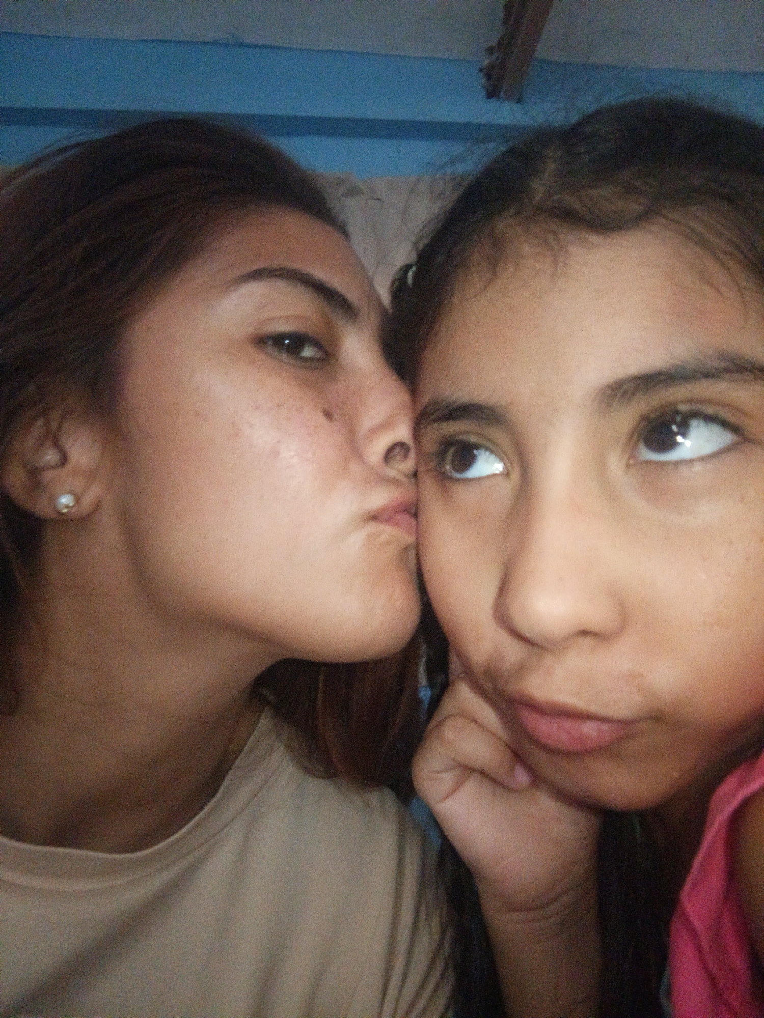 The image is a close-up, candid selfie of two females. The subject on the left, partially obscured, appears to be leaning in to kiss the subject on the right. Both individuals have brown hair and brown eyes. The subject on the left is wearing a light-colored t-shirt and a pearl earring. The subject on the right is wearing a pink top and resting her chin on her hand. The background appears to be indoors with a blue wall. The lighting suggests it is daytime. The image captures an intimate moment between the two individuals, with a sense of affection conveyed through the gesture. There is no visible text in the image.