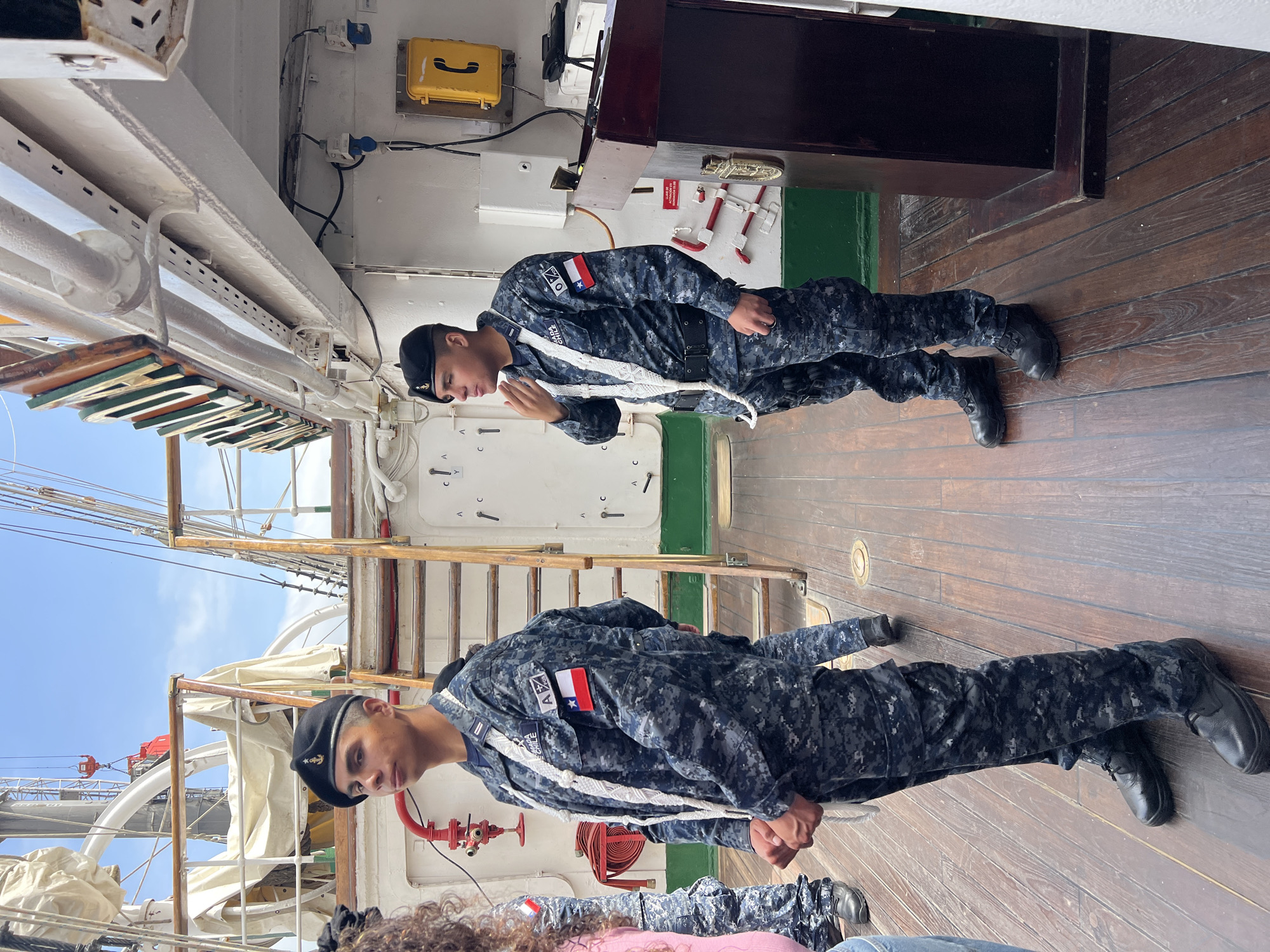 Two navy cadets stand on the wooden deck of a tall ship. They wear navy camouflage uniforms with Chilean flag patches, white belts, and black berets. The ship's rigging and masts are visible, suggesting it is either docked or sailing. The location is likely a port, possibly in Arica, Chile, given the cadets' uniforms. The environment indicates it's a sunny day with a clear sky. One cadet seems to be saluting or in a position of attention.